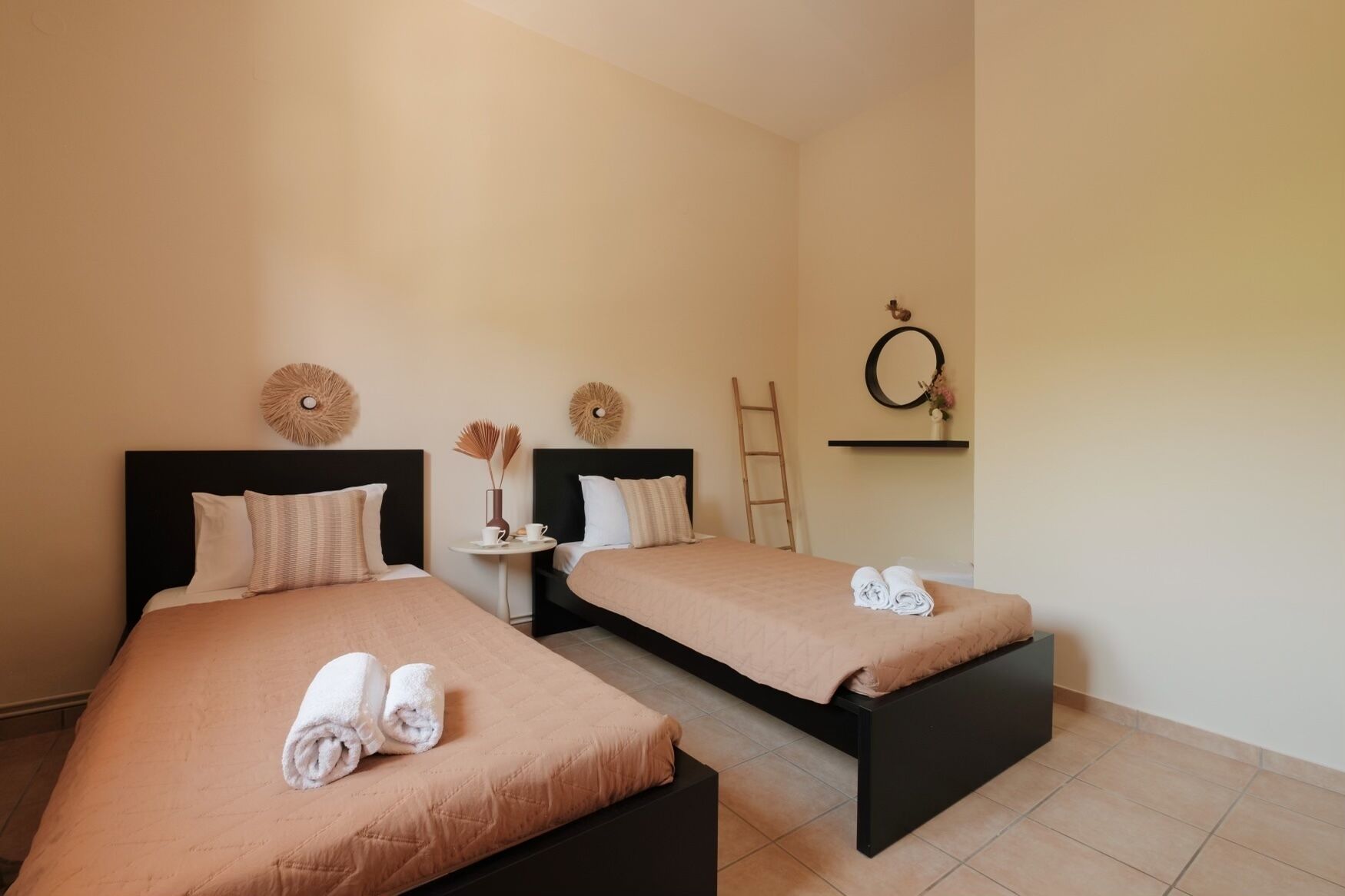 Habitación de hotel con dos camas, cada una con estructura negra y ropa de cama color beige. Hay una pequeña mesa redonda con un florero y dos tazas, y una escalera apoyada en la pared.