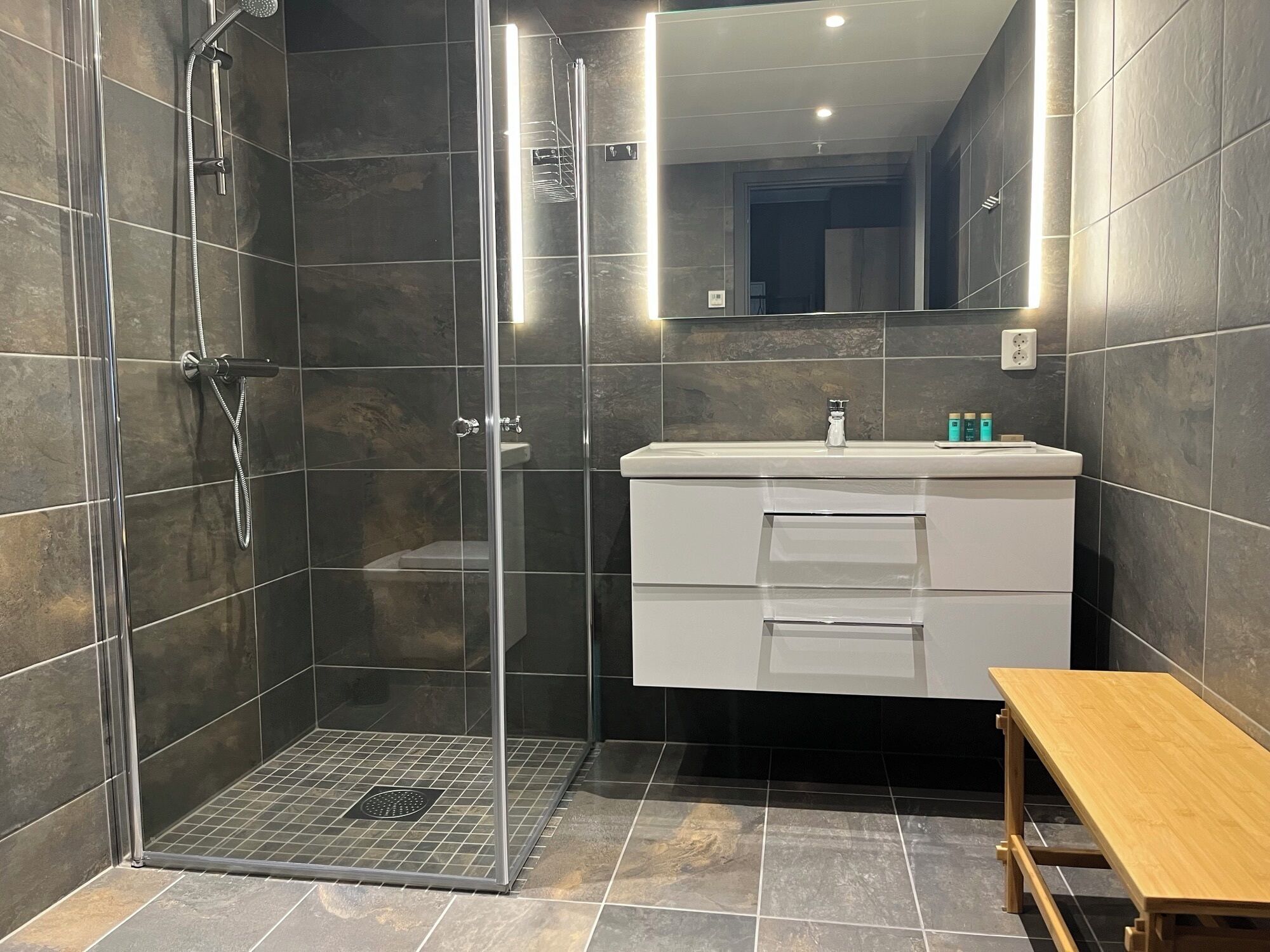 Een moderne badkamer met een douchecabine van glas, een witte wastafel met spiegel, en een houten bankje.