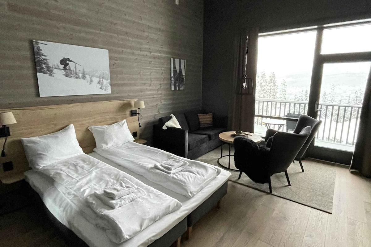 A modern hotel room with a large bed, a small sofa, a desk, and a balcony with a view.