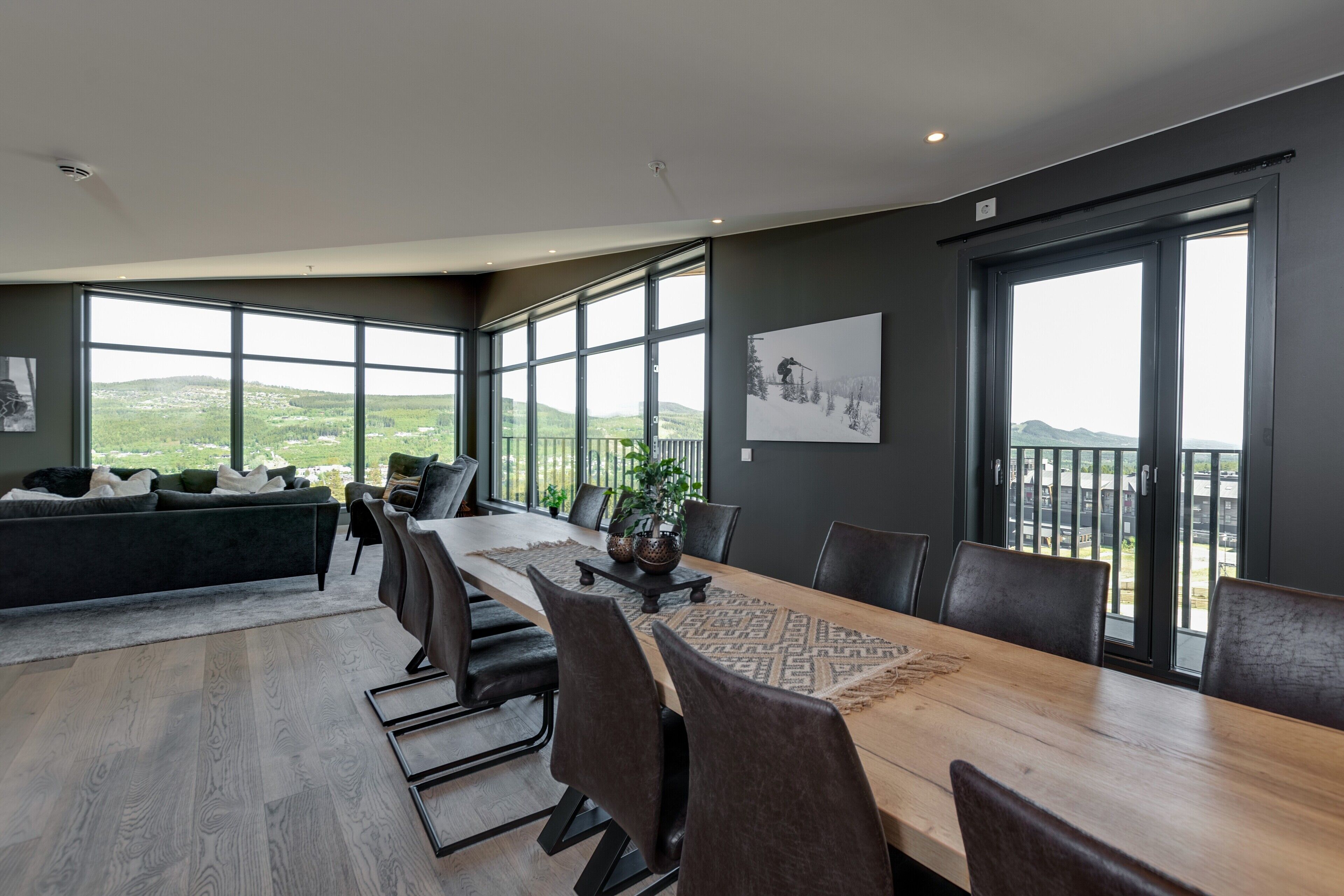A modern dining area with a long table, chairs, and a view of a green landscape through large windows.