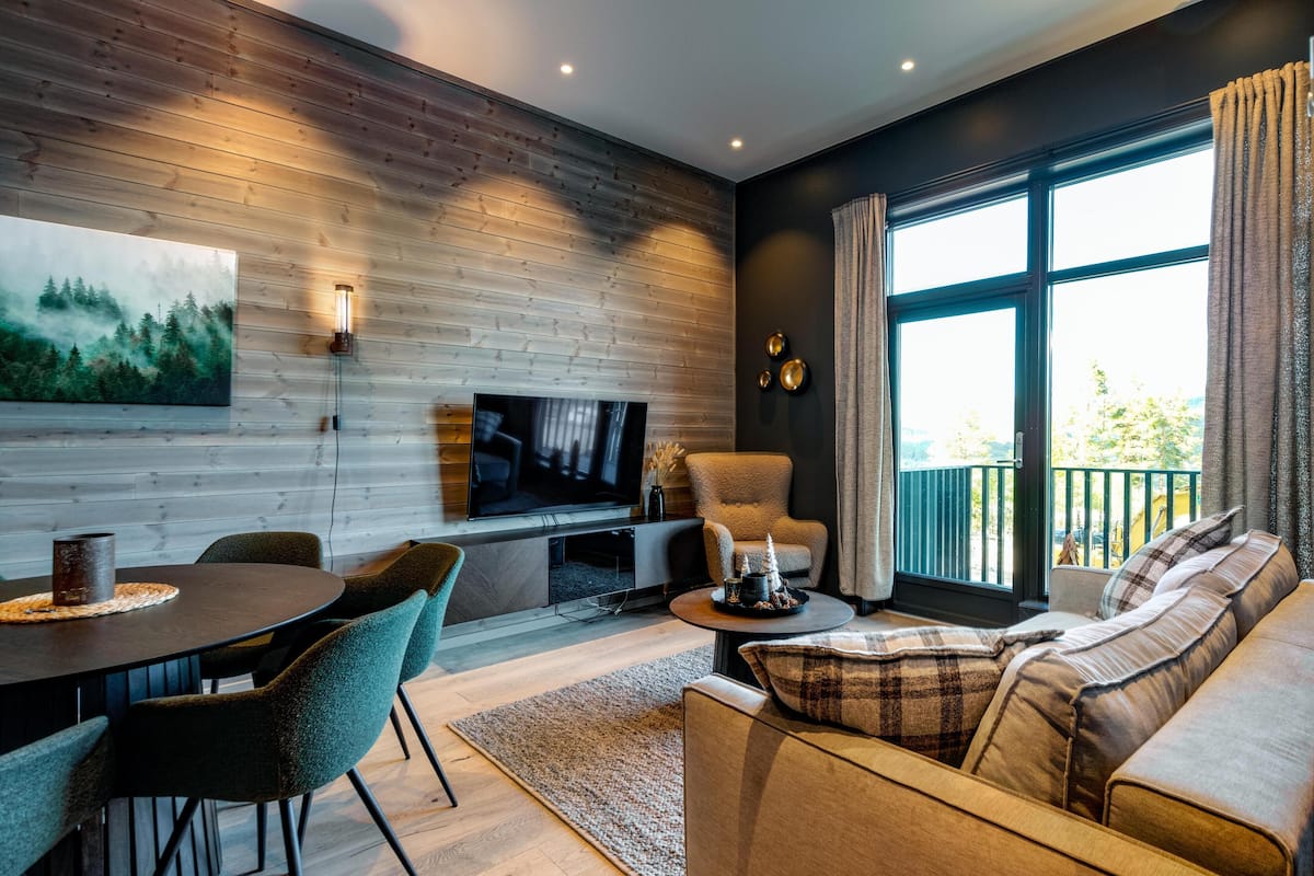 A modern living room with a wooden wall, a television, a sofa, and a dining table with chairs.