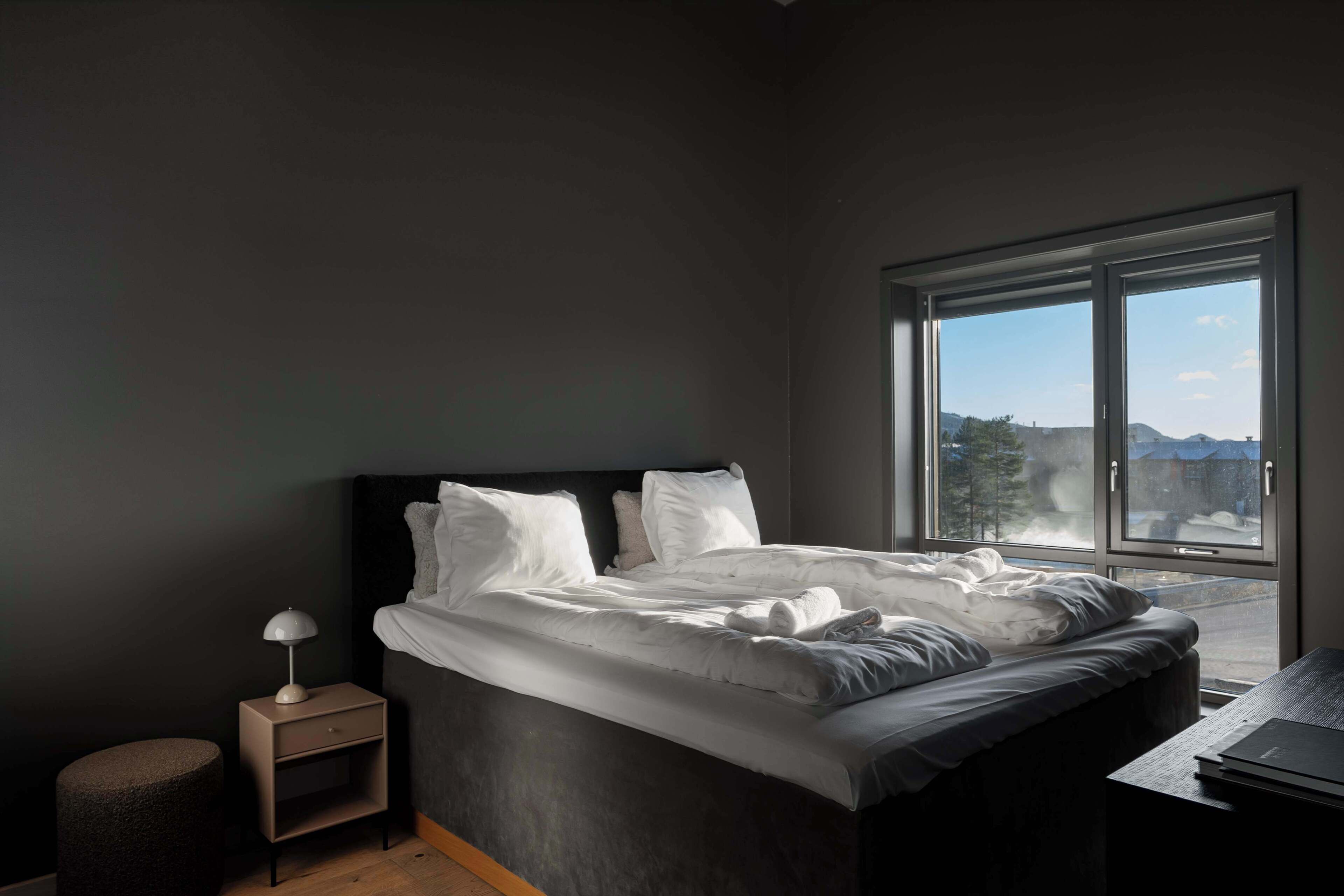 Une chambre à coucher avec un lit, une table de chevet, un petit tabouret et une grande fenêtre donnant sur un beau paysage.