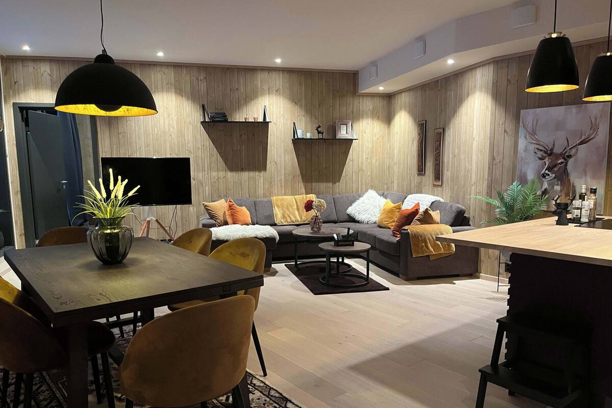A modern living room with a wooden floor, a dining table with chairs, a sofa with cushions, and pendant lighting.