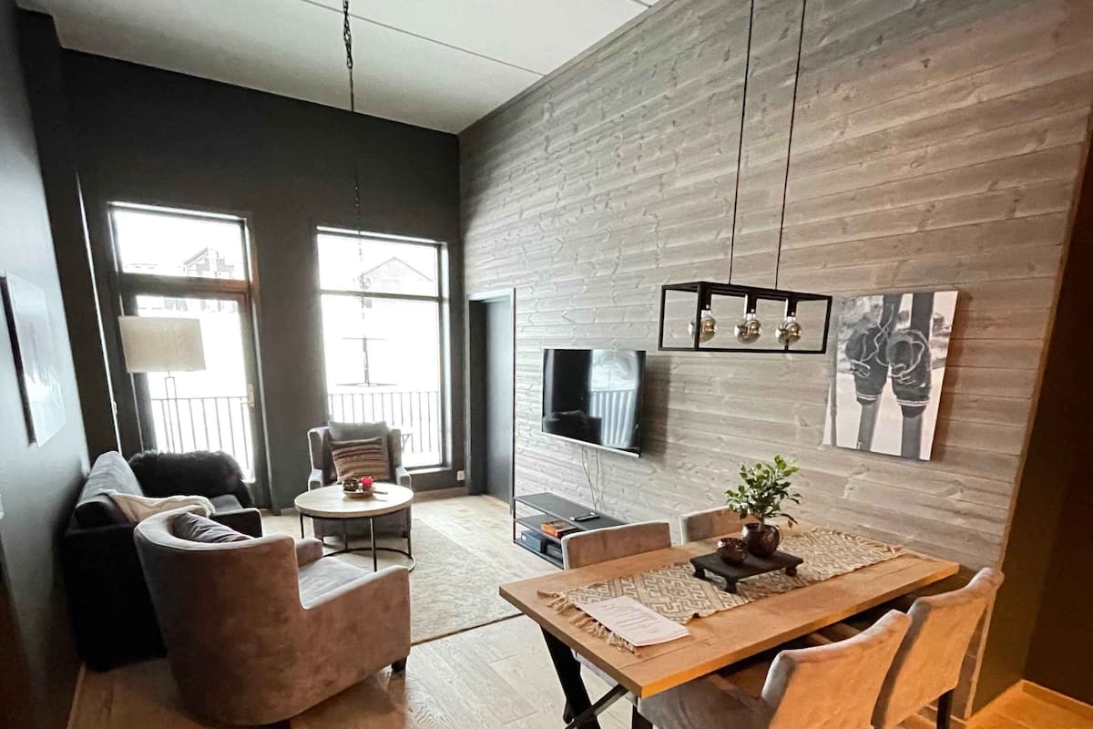 A modern living room with a dining area, featuring a wooden floor, a sofa, a coffee table, and a TV mounted on the wall.