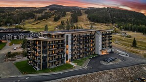 Un hotel de varios pisos con balcones, zona de estacionamiento y vistas a un paisaje montañoso.