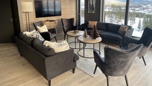 Un salón moderno con un sofá gris oscuro, dos sillones, una mesa de centro y un televisor montado en la pared. Amplias ventanas ofrecen vistas a un paisaje nevado.