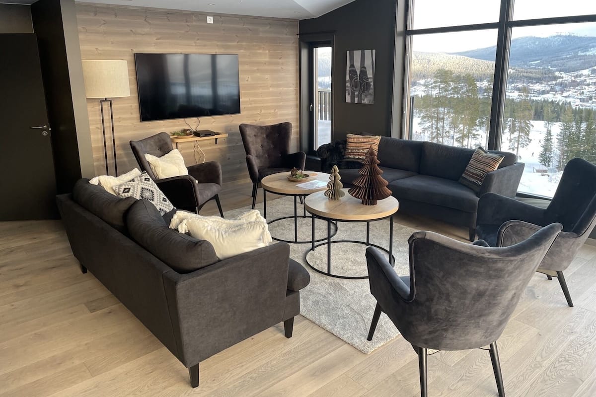 A modern living room with a dark grey sofa, two armchairs, a coffee table, and a TV mounted on the wall. Large windows offer a view of a snowy landscape.
