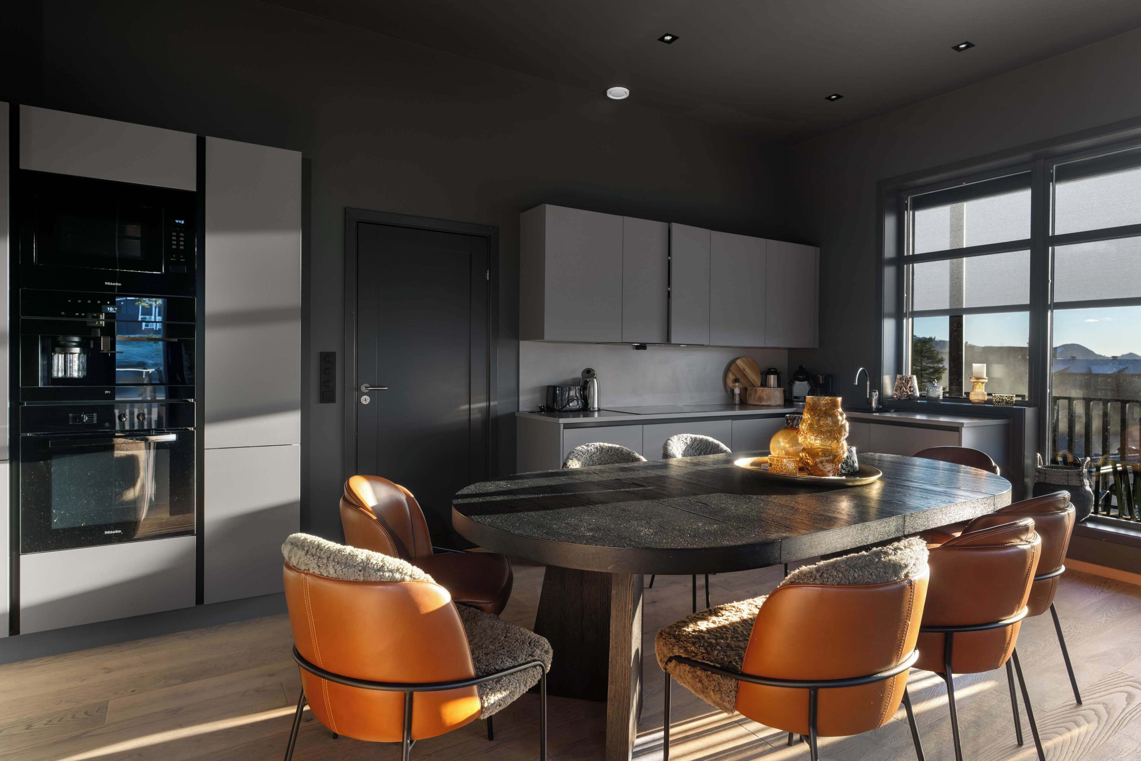 A modern kitchen with a built-in oven and microwave, a dining area with a round table and leather chairs, and large windows offering a view of the mountains.