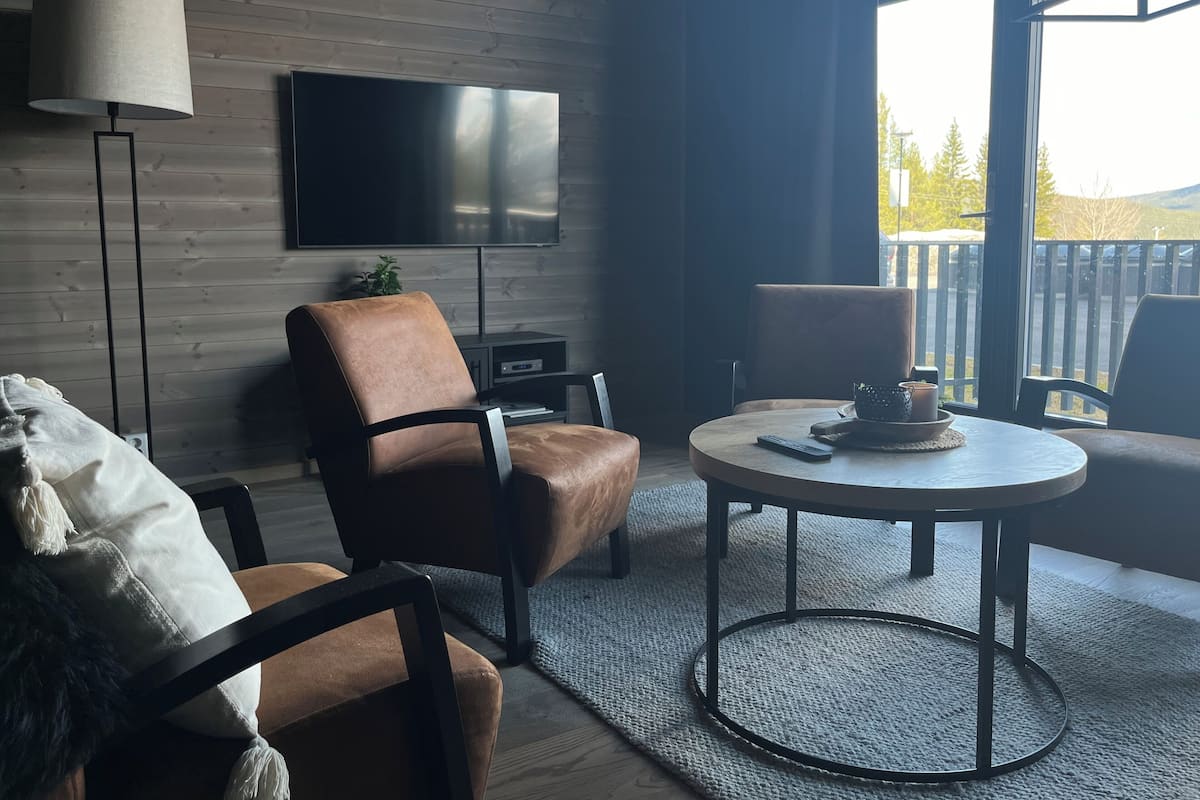 A modern living room with a leather armchair, a round coffee table, a flat-screen TV mounted on a wooden wall, and a view of a deck through a large window.
