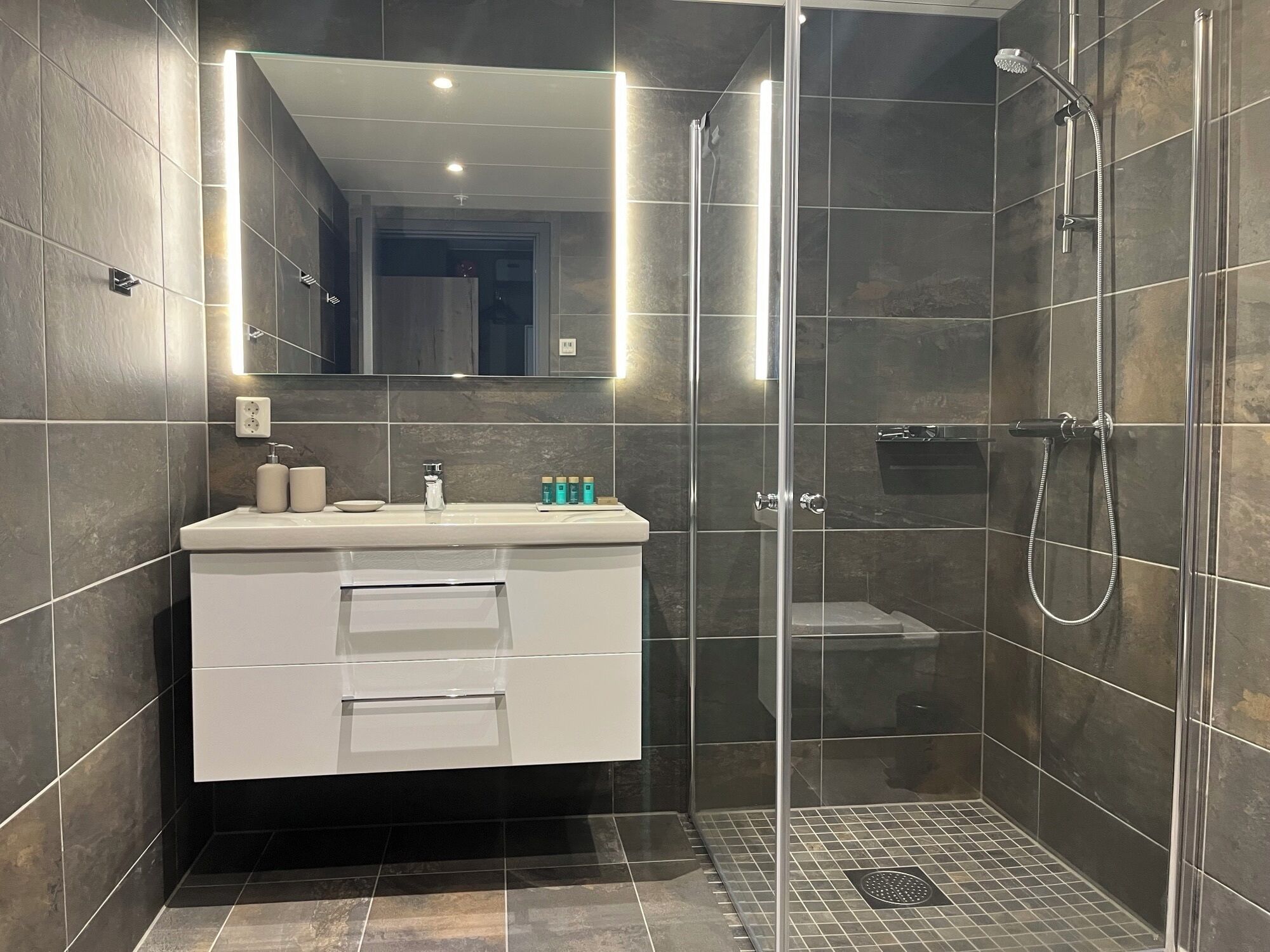 Un baño moderno con ducha acristalada, tocador blanco con espejo y paredes de azulejos oscuros.