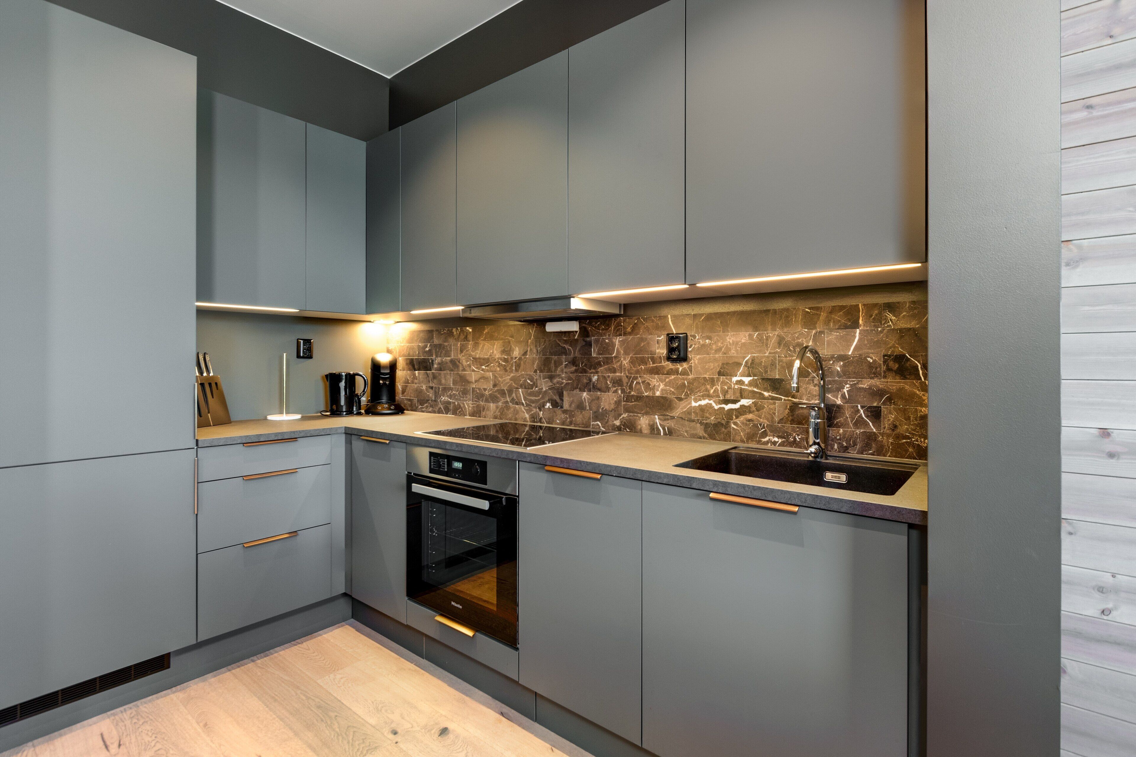 Una cocina moderna con gabinetes grises, horno empotrado, protector contra salpicaduras de mármol y un lavamanos de acero inoxidable.