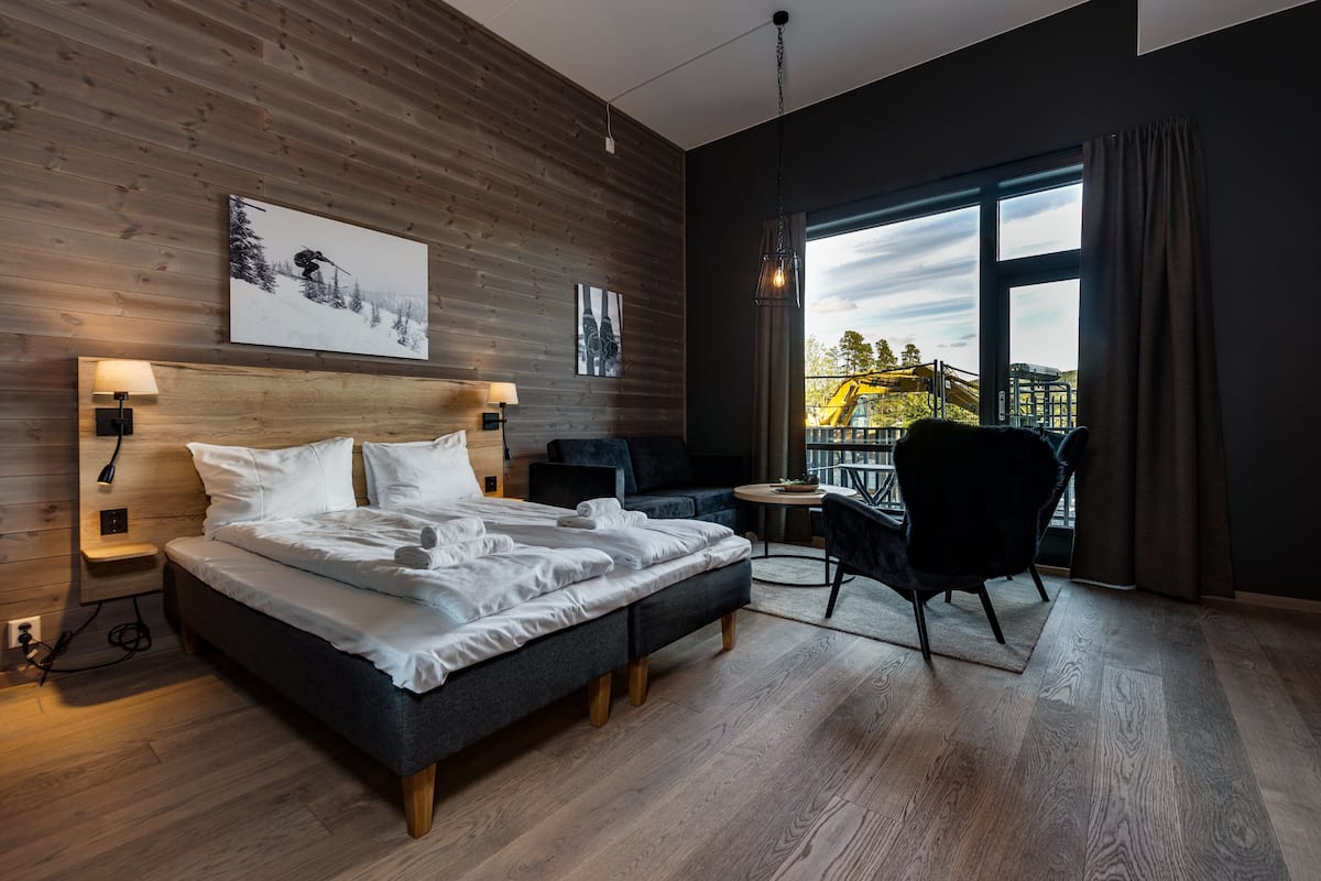 A modern hotel room with a wooden wall, a large bed, a small desk, a chair, and a balcony with a view of construction.