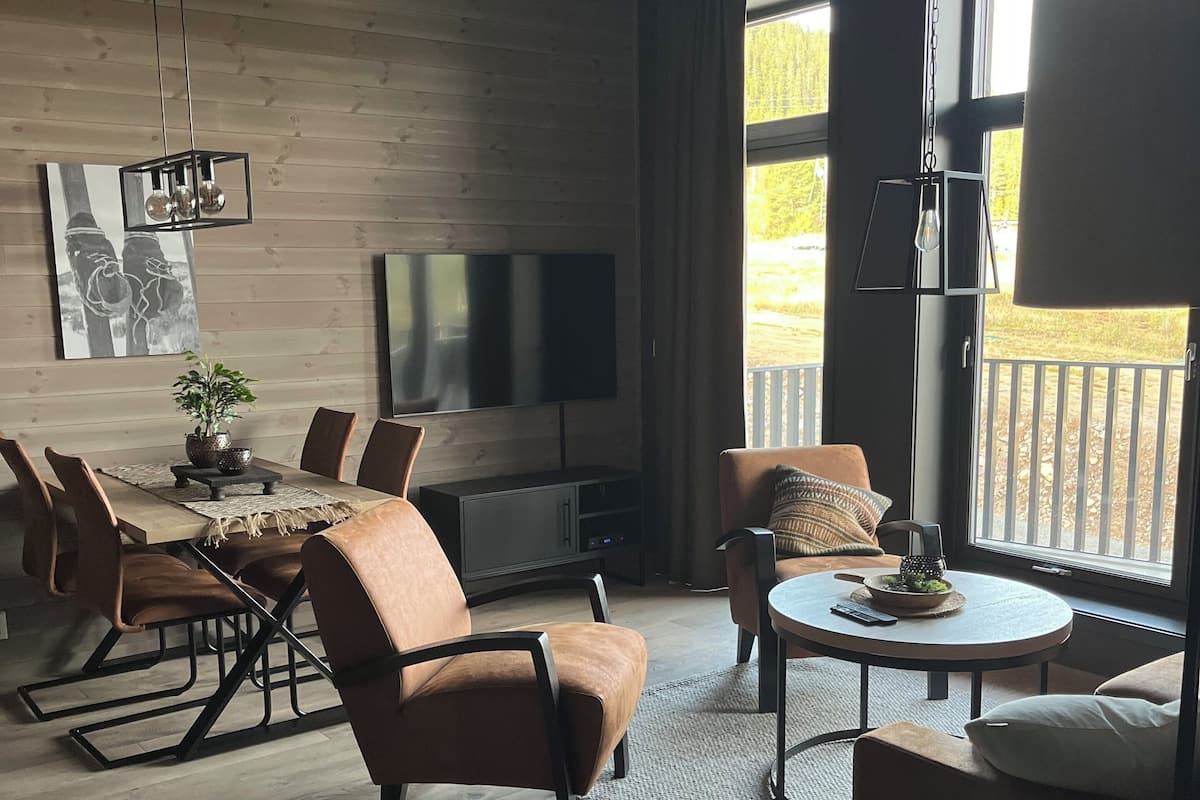 A modern living room with a dining area, wooden walls, a flat-screen TV, and large windows.