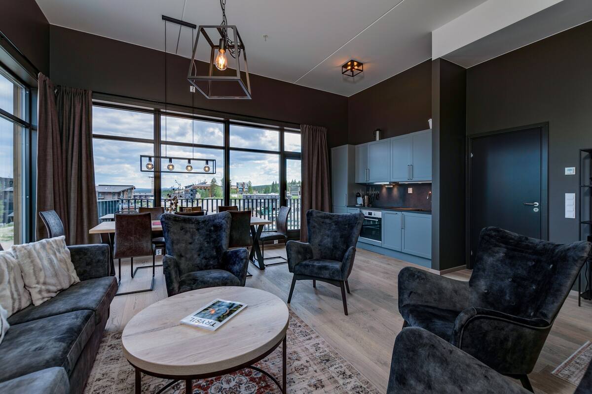 A modern living room with a round coffee table, dark armchairs, and a dining area with a table and chairs. A large window offers a view of the outdoors.