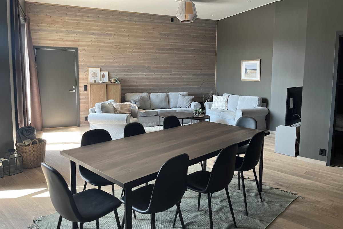 A modern living room with a wooden wall, a grey sofa set, a dining table with chairs, and a television.