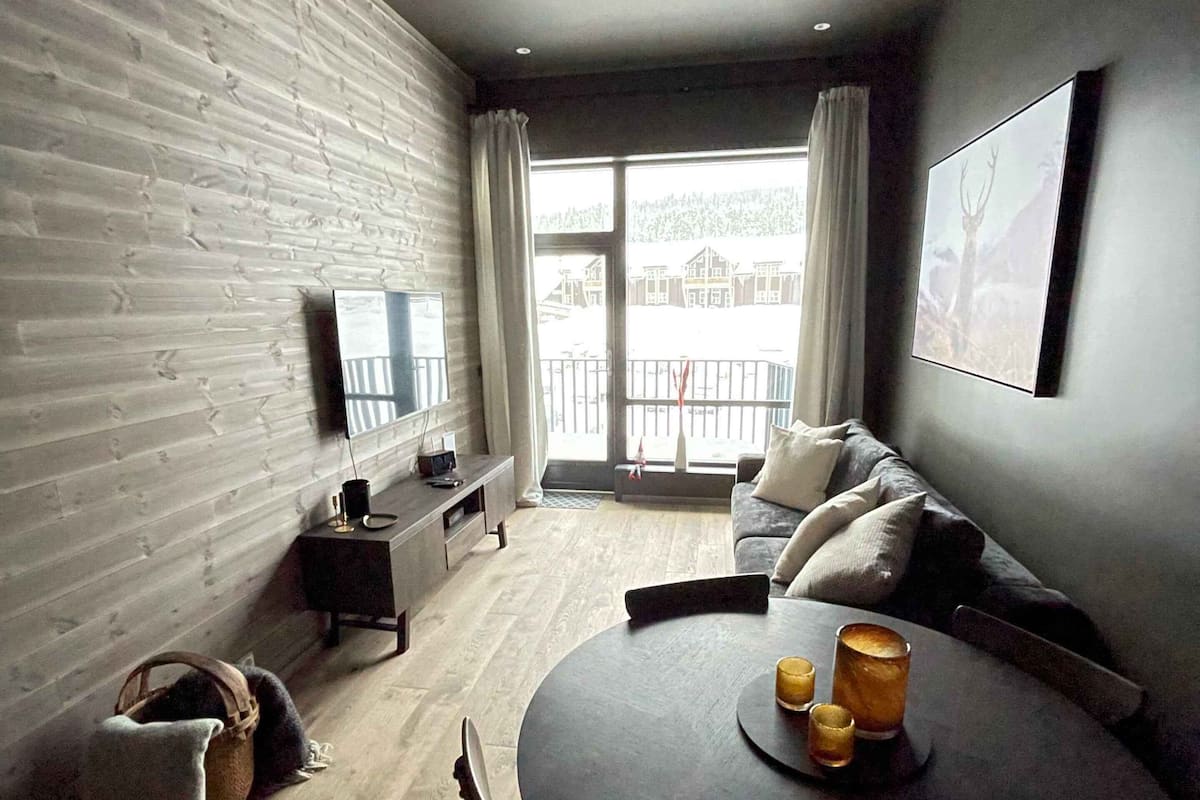 A modern living room with a round dining table, a sofa, a TV, and a large window with a view of houses.