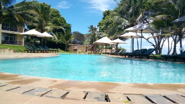 Ein Swimmingpool mit Liegestühlen und Sonnenschirmen, umgeben von Palmen und blauem Himmel.