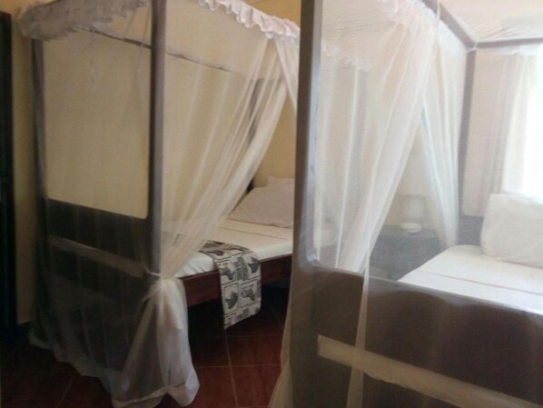 A room with a bed under a white canopy, a bedside table with a patterned cloth, and a window with curtains. - Coral Beach Resort (Diani Beach)