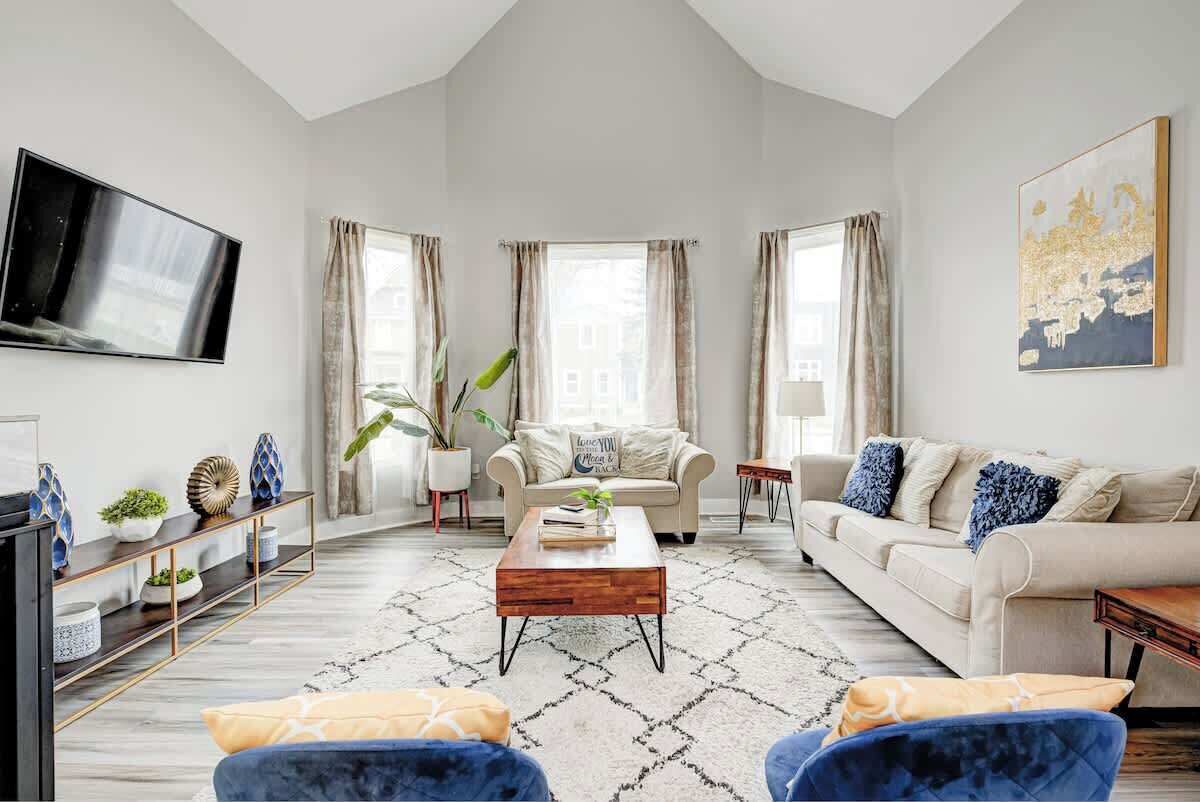 Een moderne woonkamer met een hoge plafond, een flatscreen televisie aan de muur, een beige bank, een blauwe fauteuil, een houten salontafel en een kamerkleed met een patroon.