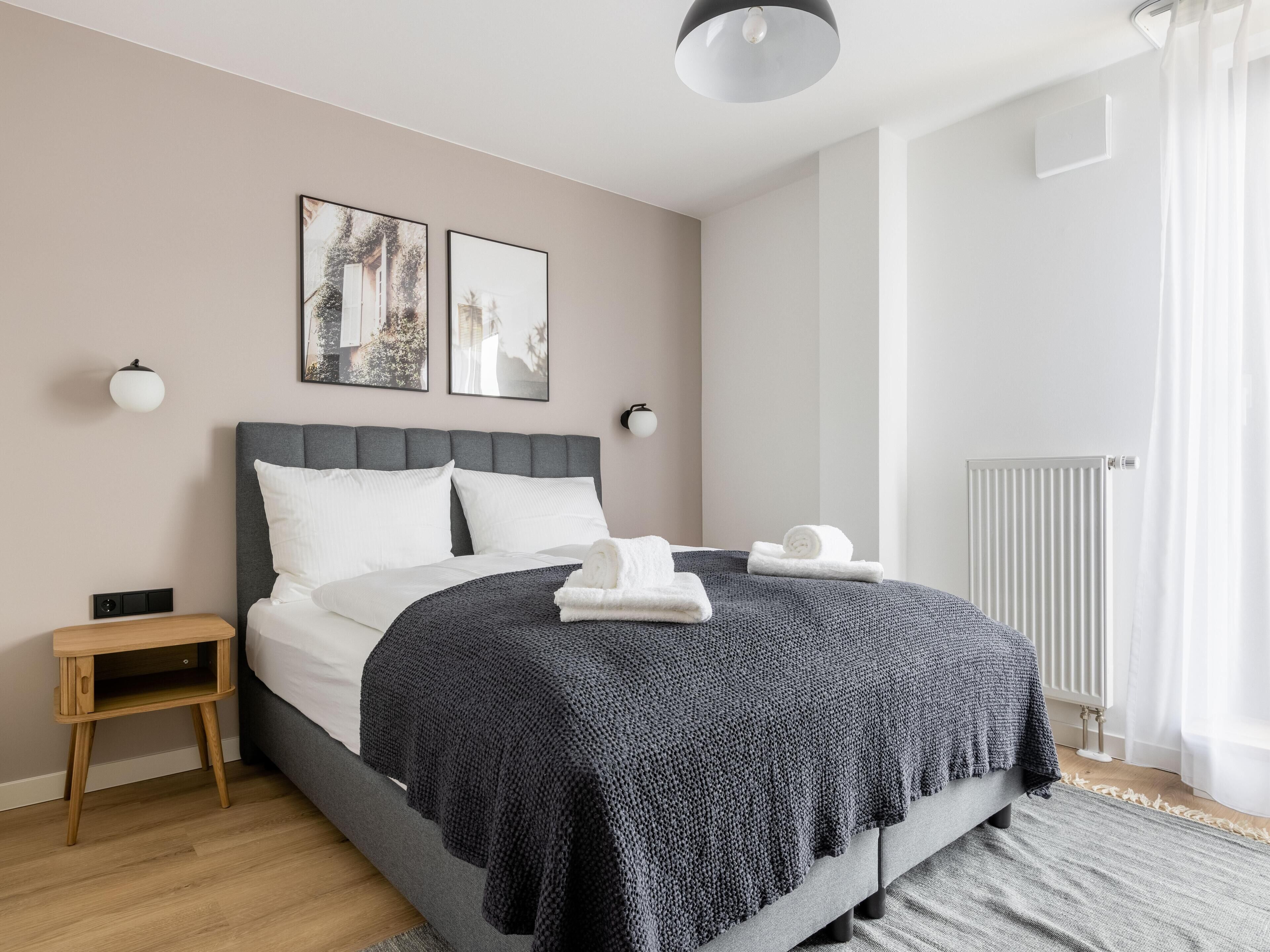 Een moderne slaapkamer met een grijs bed, witte kussens, een donkere gebreide plaid, een klein houten nachtkastje en twee ingelijste foto’s aan de muur.
