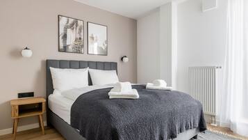 Een moderne slaapkamer met een grijs bed, witte kussens, een donkere gebreide plaid, een klein houten nachtkastje en twee ingelijste foto’s aan de muur.