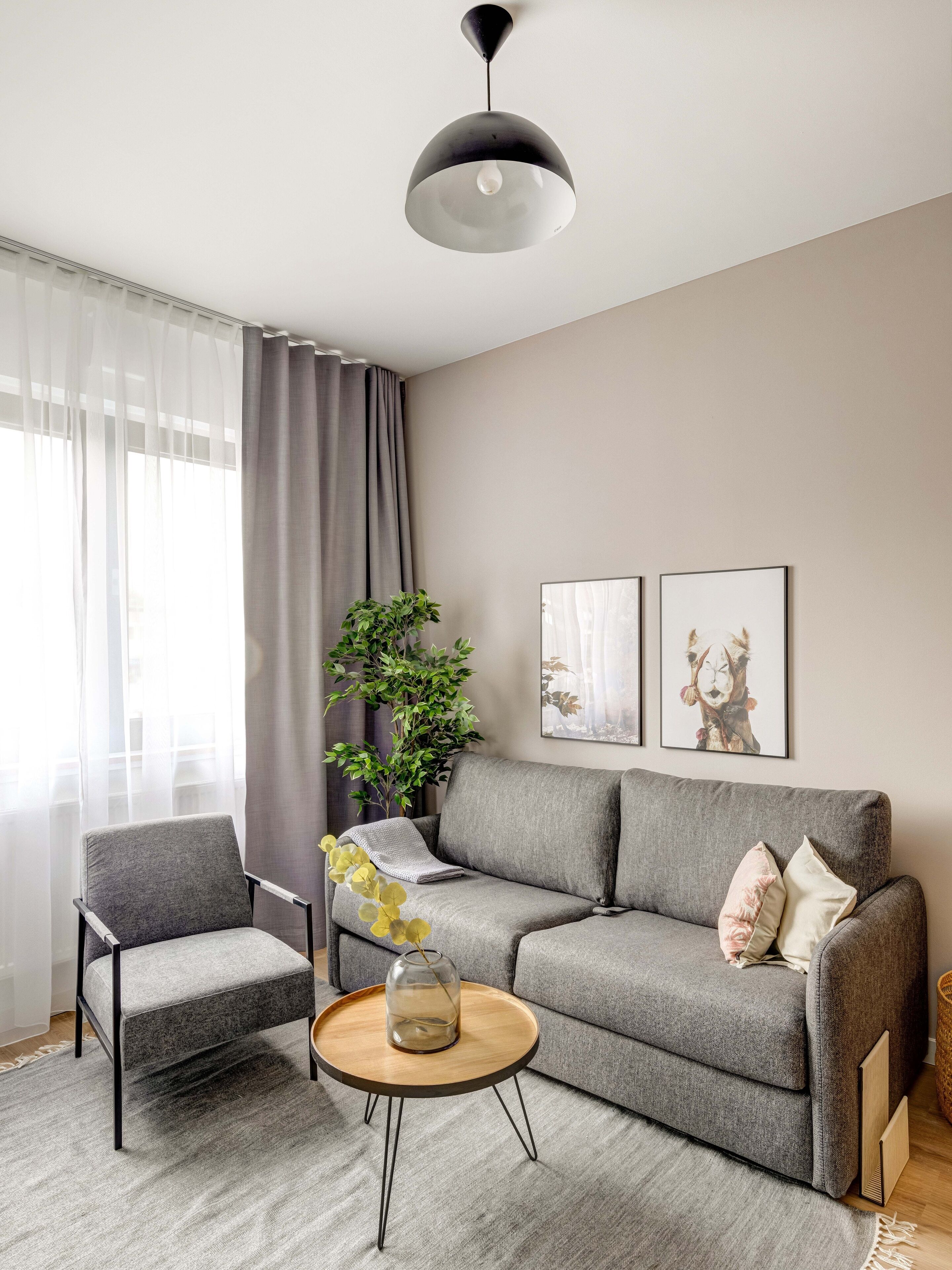Een moderne woonkamer met een grijze bank, een klein houten bijzettafeltje, een grijze fauteuil en een groot raam met lichte gordijnen.