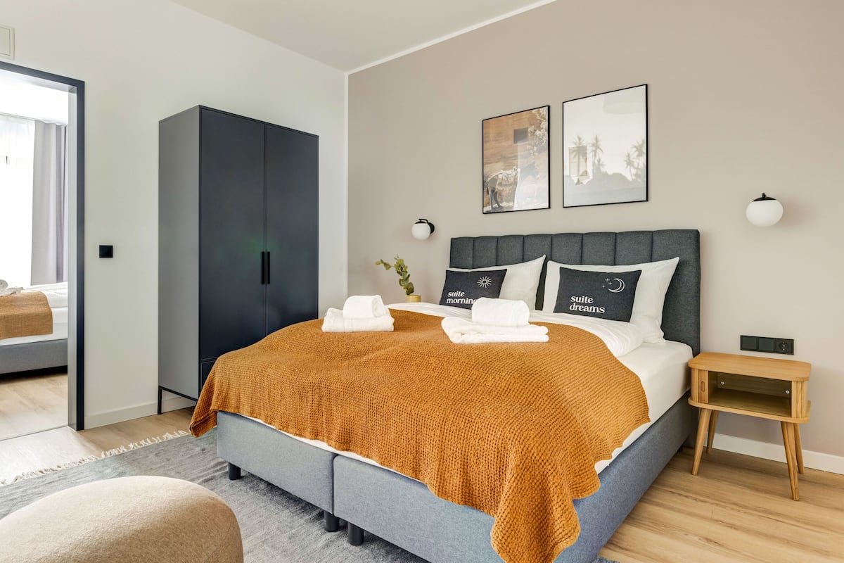 A modern bedroom with a large bed, a dark wardrobe, and a small bedside table.