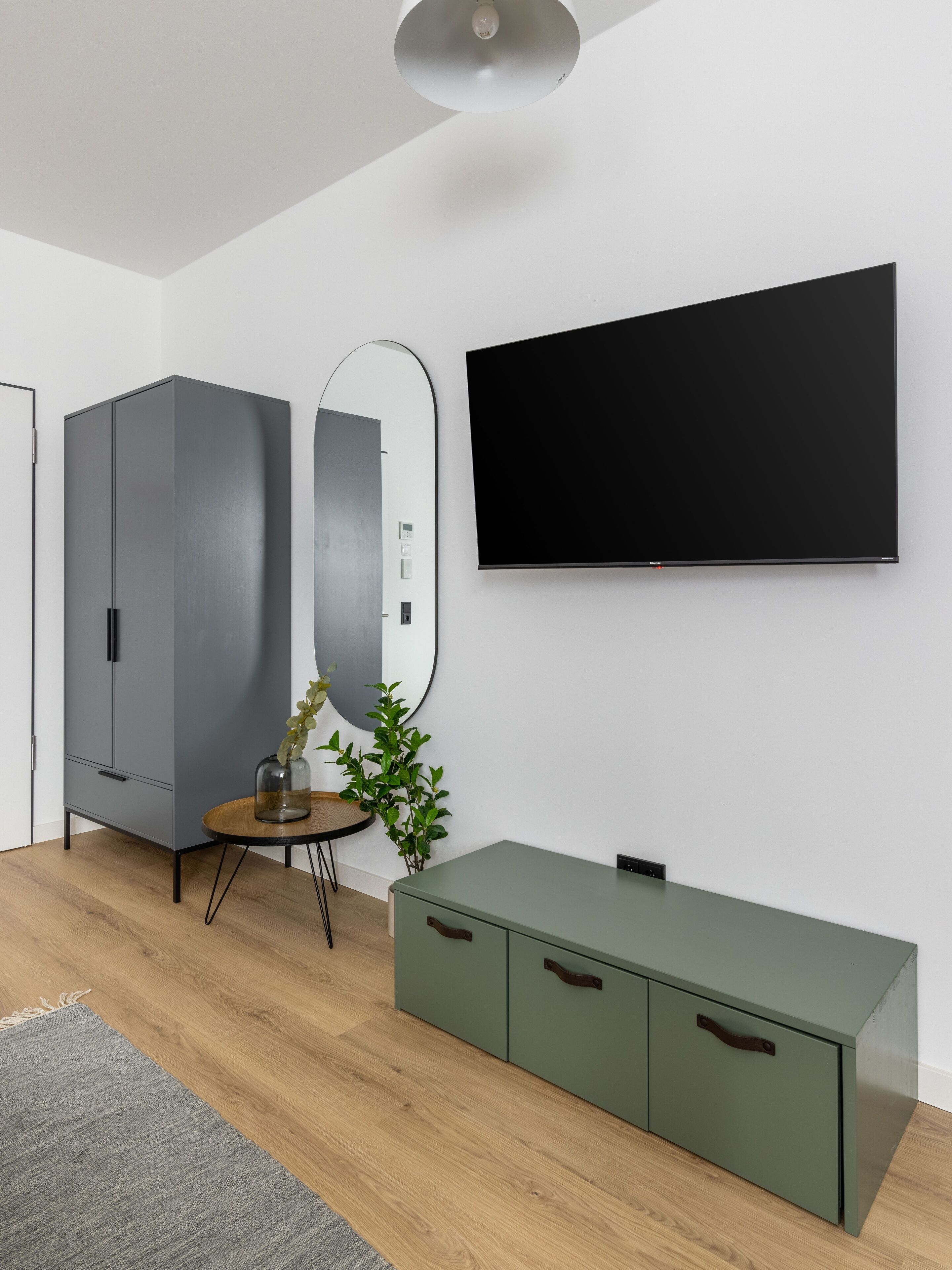 A modern room with a grey wardrobe, a round mirror, a green cabinet, and a flat-screen TV mounted on the wall.