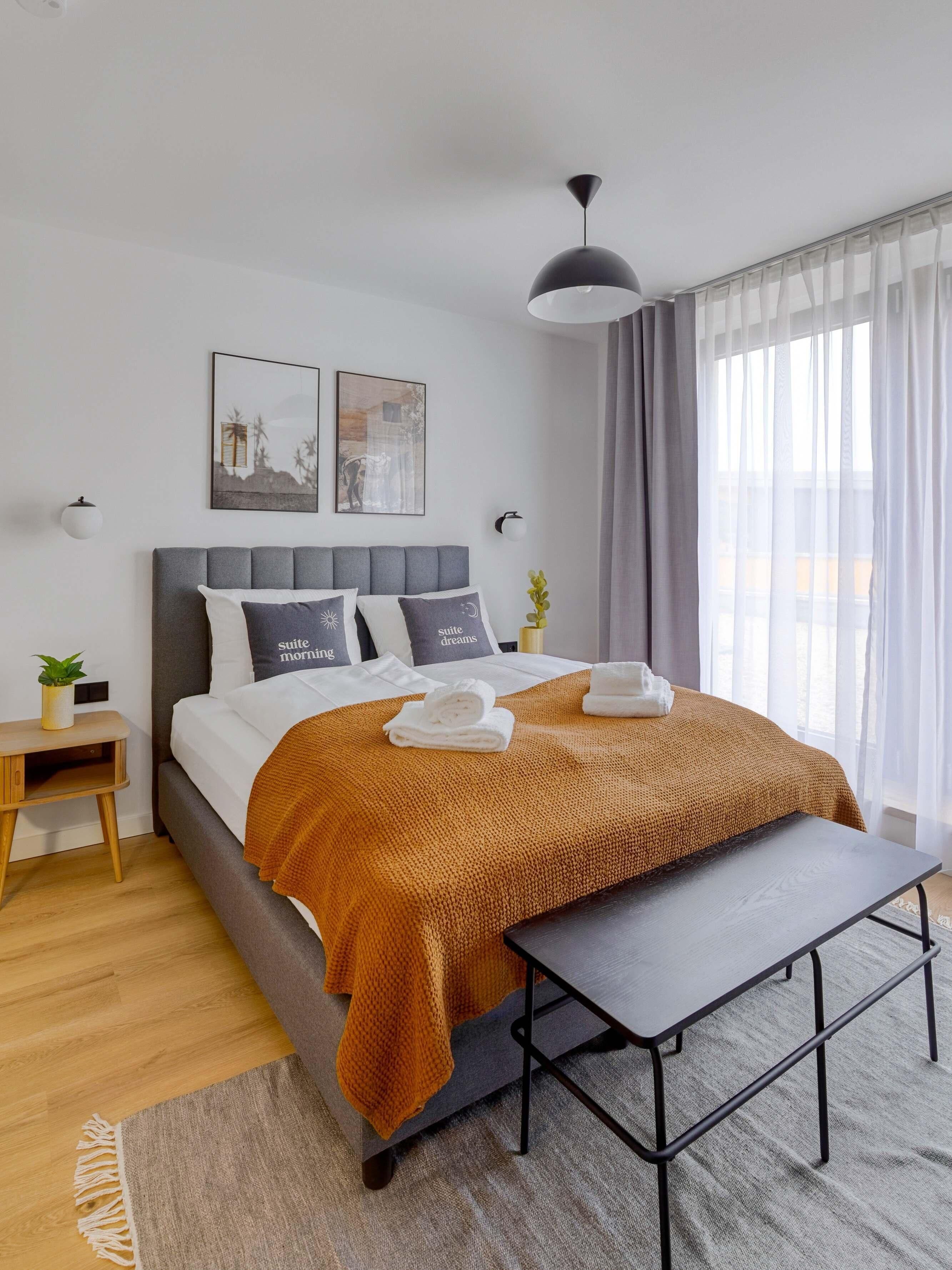 Un dormitorio moderno con una cama tapizada en gris, una mesita de noche de madera, una pequeña mesa y un ventanal con cortinas traslúcidas.