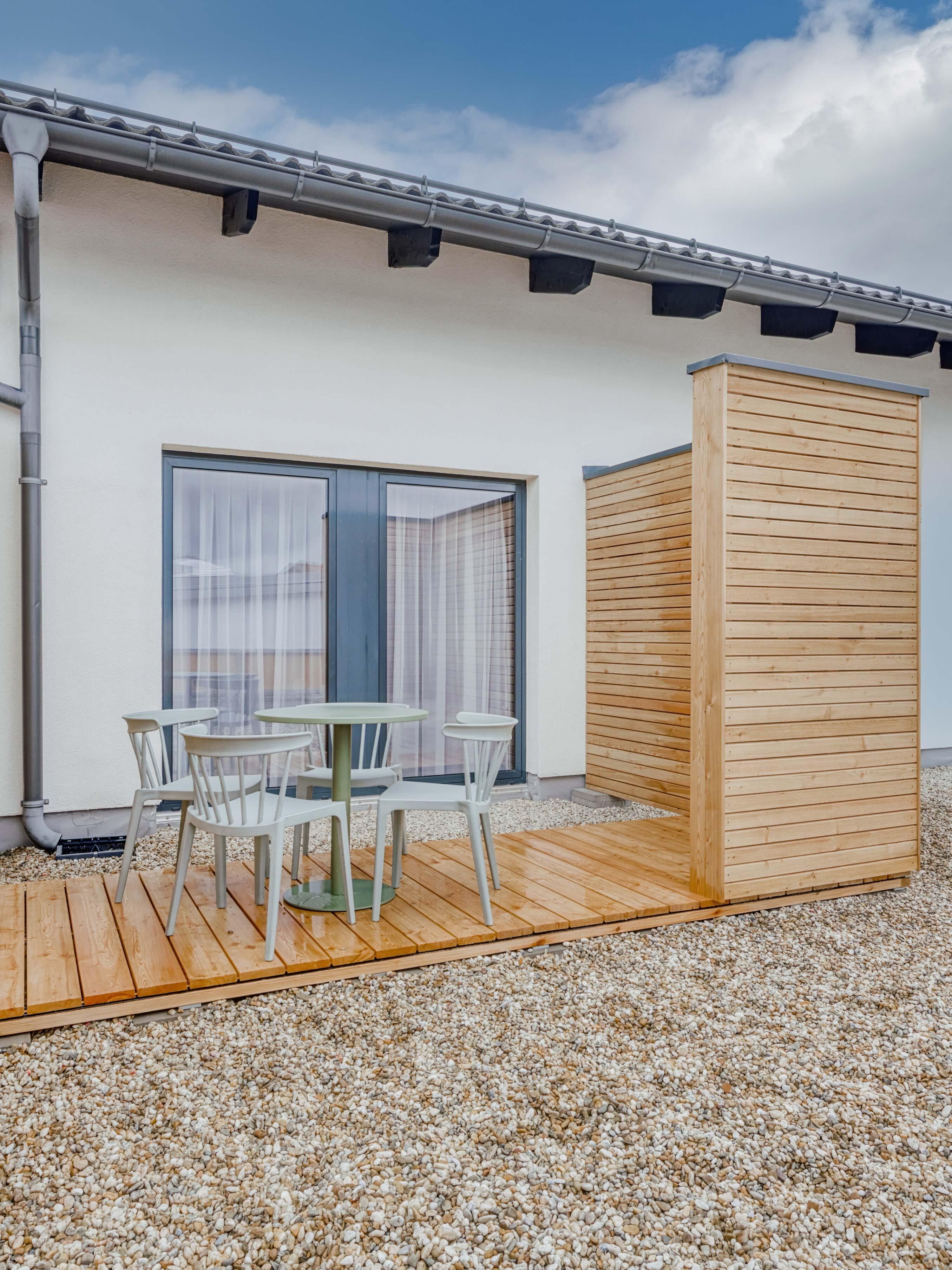 A modern outdoor seating area with a wooden deck, a table with chairs, and a large window with curtains.
