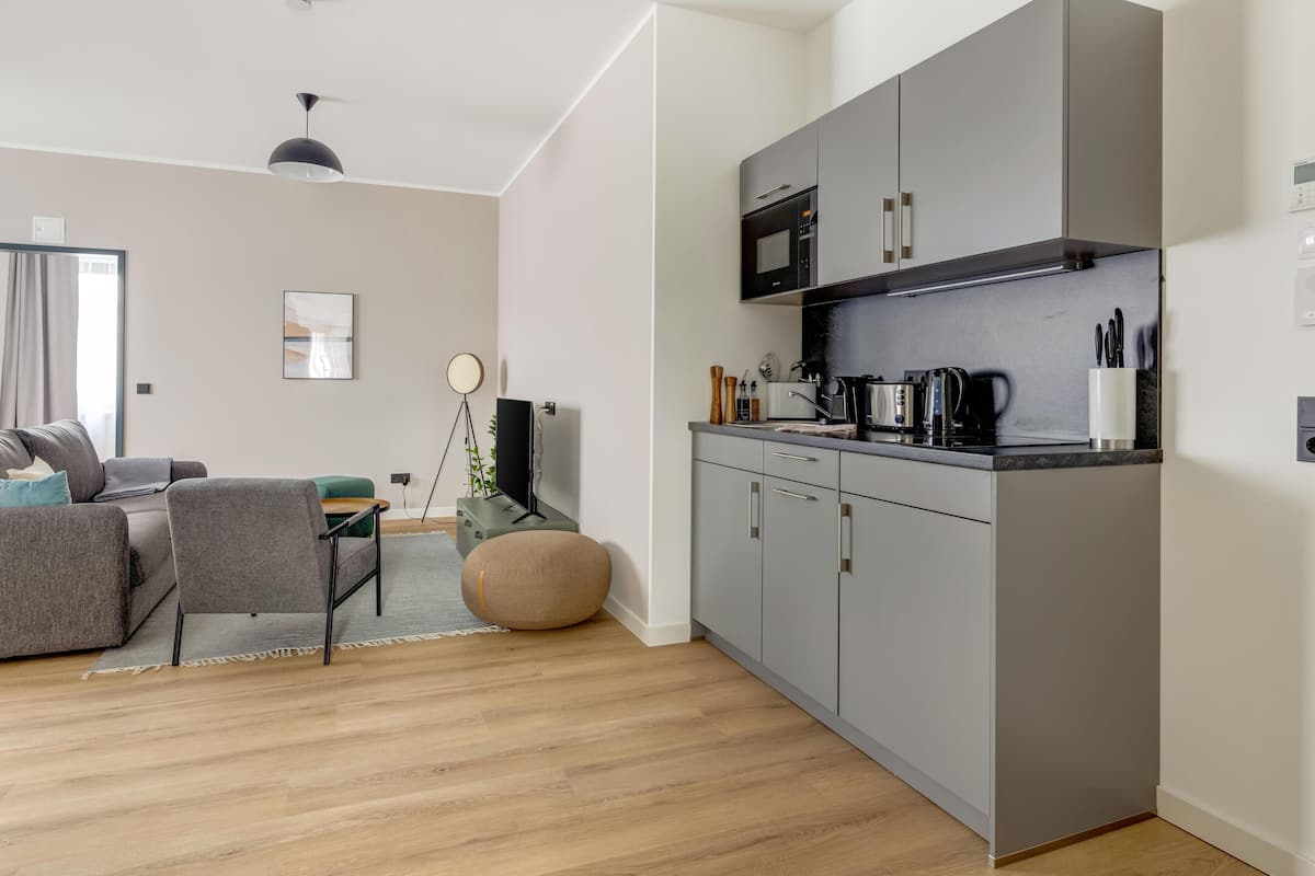 A modern kitchen with grey cabinets, a black countertop, and a coffee machine. There is a dining area with a grey sofa and a round table.