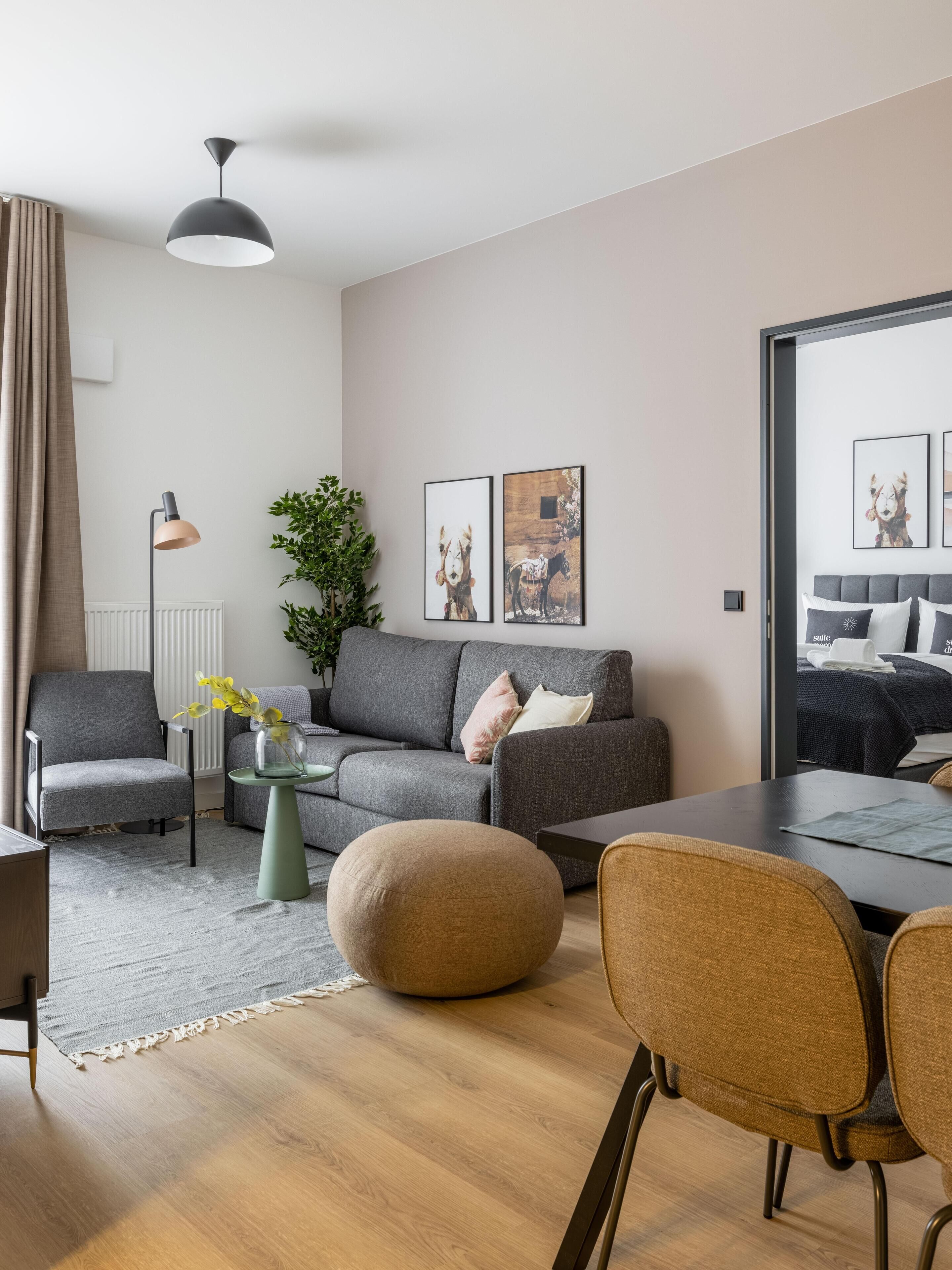 Una sala moderna con un sofá gris, un otoman redondo, una mesa de comedor con sillas, una lámpara de pie y dos cuadros enmarcados en la pared.
