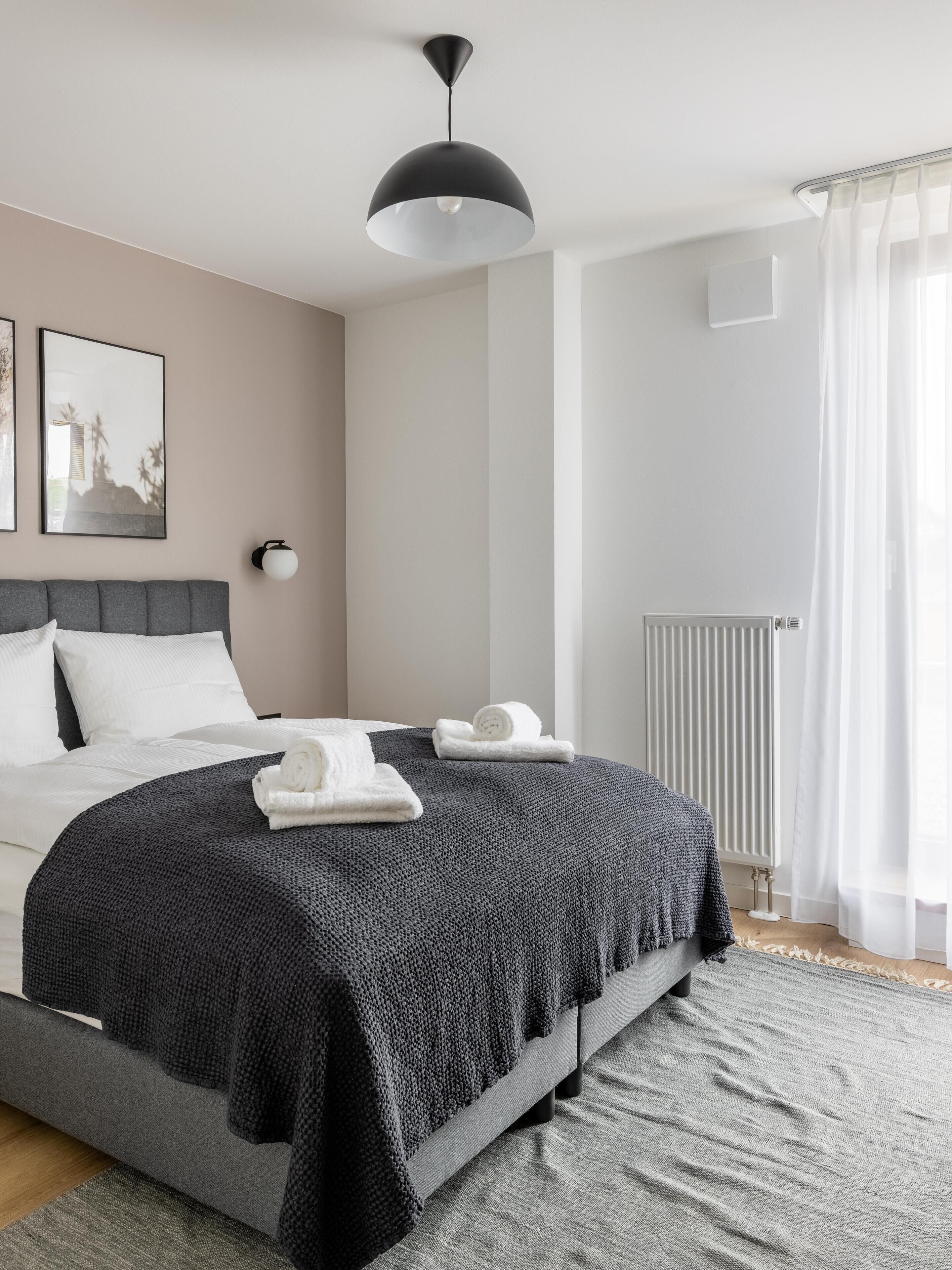Ein modernes Schlafzimmer mit einem Bett, einer schwarzen Pendelleuchte, einer Heizung und einem Fenster mit leichtem Vorhang.