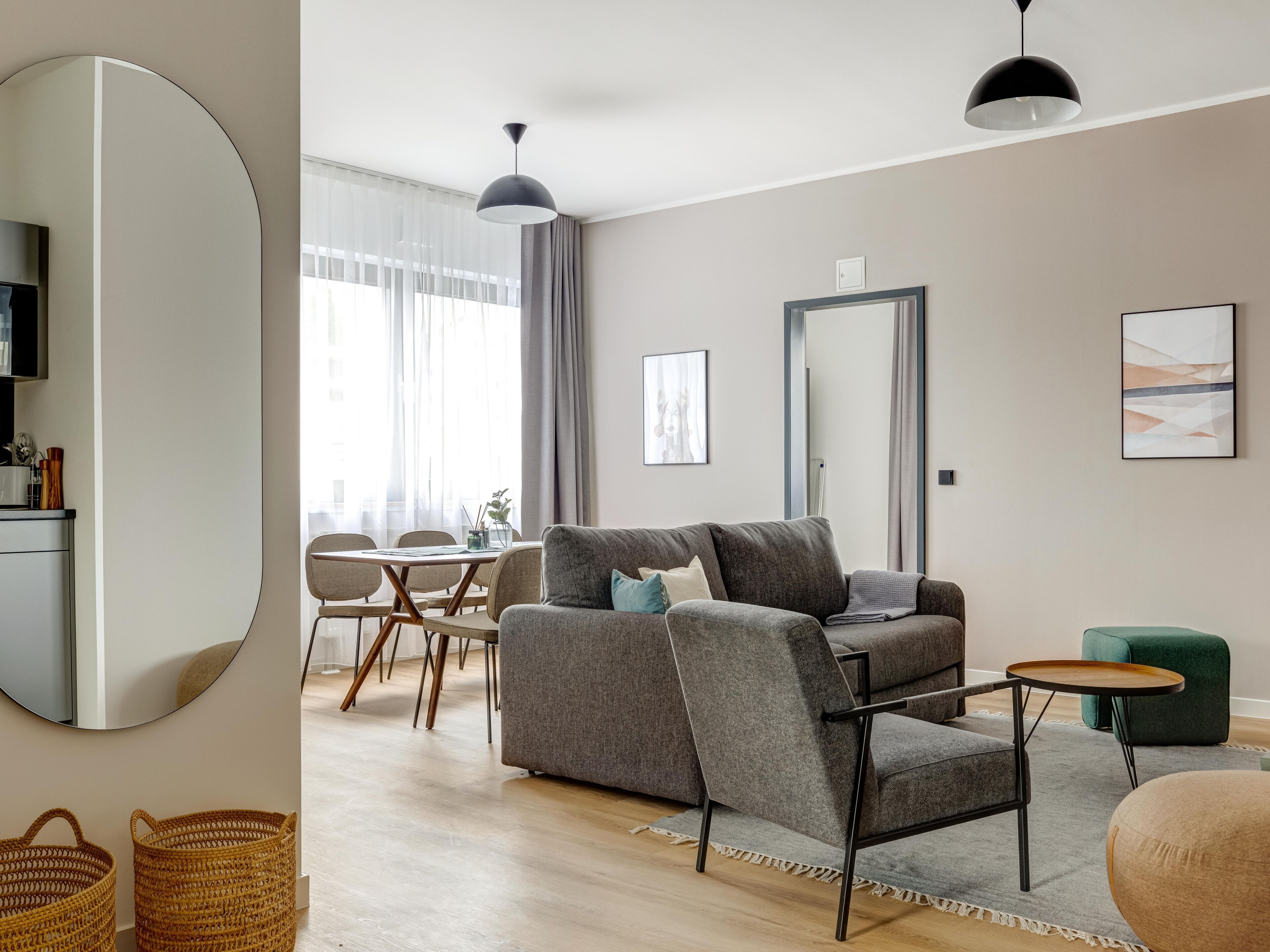 A modern living room with a grey sofa, a small round table, and a green armchair. A dining area with a wooden table and chairs is visible in the background.
