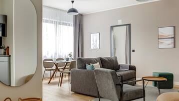 A modern living room with a grey sofa, a small round table, and a green armchair. A dining area with a wooden table and chairs is visible in the background.