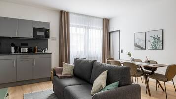 A modern living room with a grey sofa, a dining table with chairs, a kitchen area with grey cabinets, and a large window with curtains.