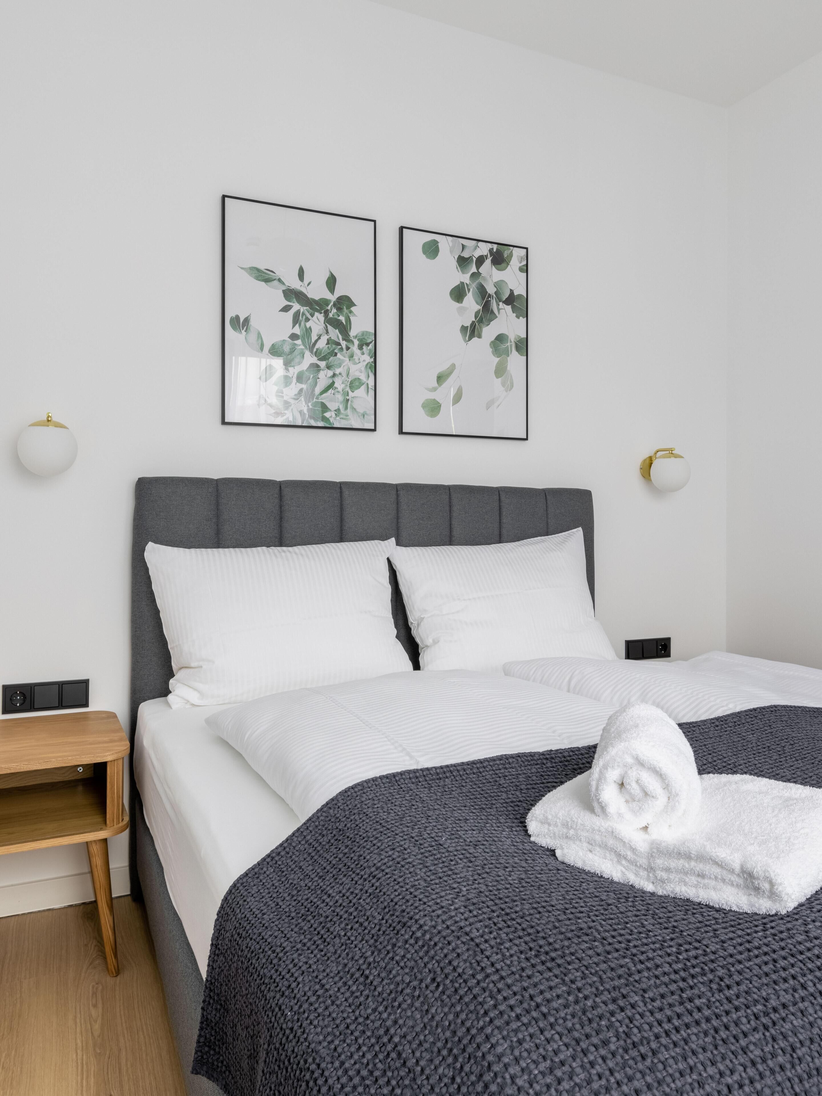 Una cama bien tendida con sábanas blancas y una manta de punto gris oscuro, una mesita de noche de madera y dos grabados botánicos enmarcados en la pared.