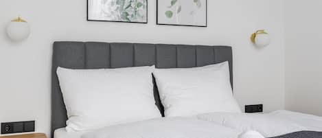 Uma cama bem arrumada com lençóis brancos e um cobertor tricotado cinza escuro, um criado-mudo de madeira e duas gravuras botânicas emolduradas na parede.