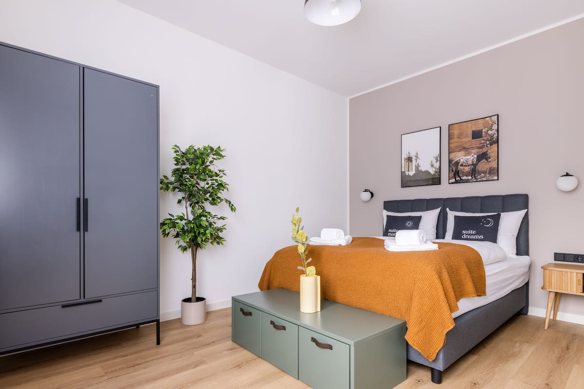 A modern bedroom with a grey wardrobe, a green bedside table, a bed with a mustard blanket, a potted plant, and two wall-mounted lights.