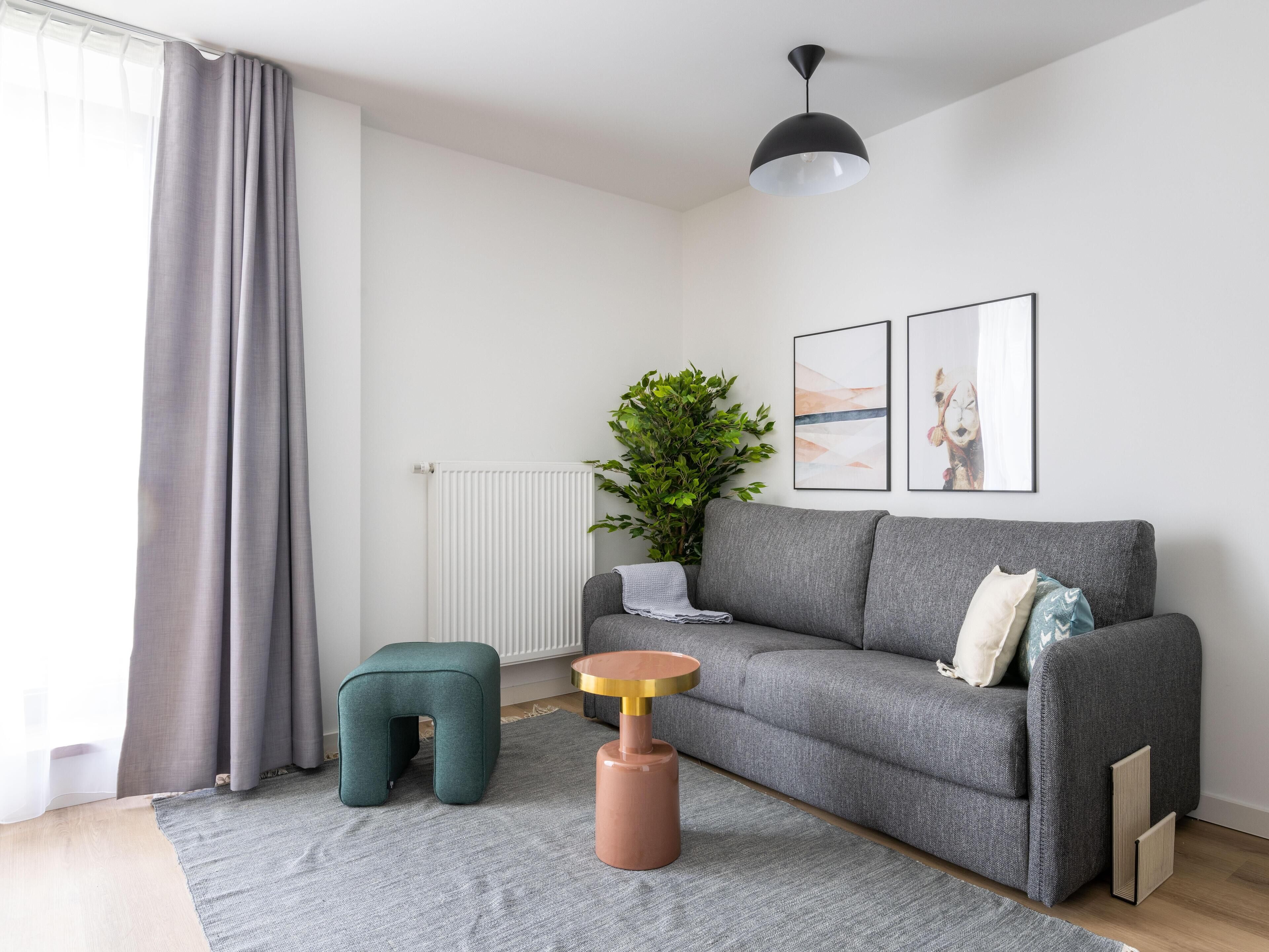 Ein modernes Wohnzimmer mit einem grauen Sofa, einem kleinen grünen Hocker, einem runden Holztisch und einem Blumentopf.