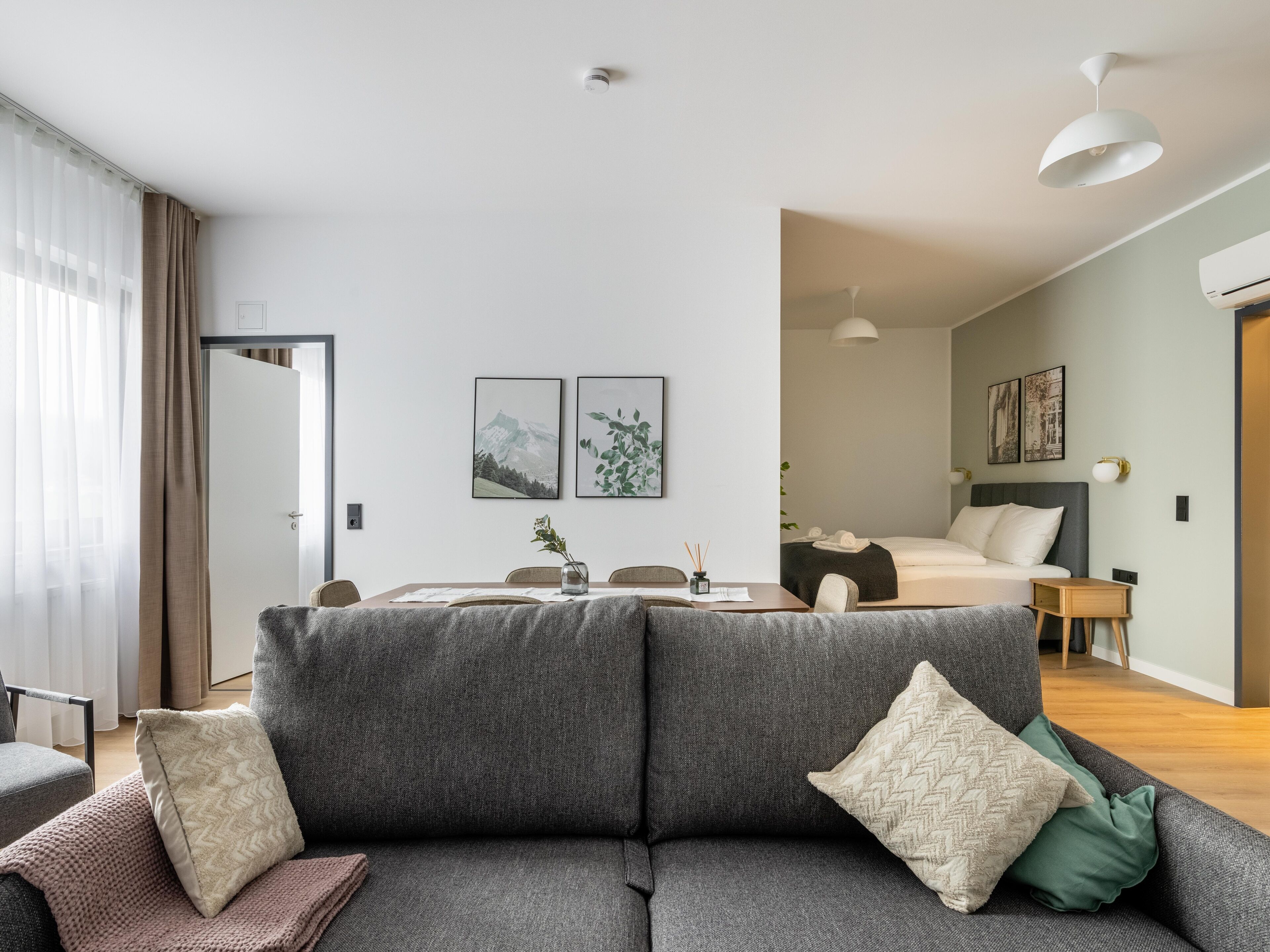 Une chambre d’hôtel moderne avec un canapé gris, une table à manger et un lit doté d’une tête de lit grise. On y trouve deux tableaux encadrés accrochés au mur, une petite table avec un vase et une table de chevet avec une lampe.