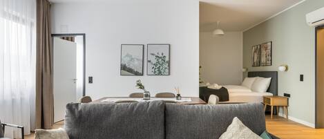 Una habitación de hotel moderna con un sofá gris, una mesa de comedor y una cama con cabecera gris. Hay dos cuadros enmarcados en la pared, una mesita con un jarrón y una mesita de noche con una lámpara.