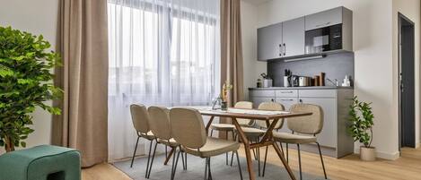 A modern dining area with a wooden table and chairs, a grey rug, and a kitchen in the background.