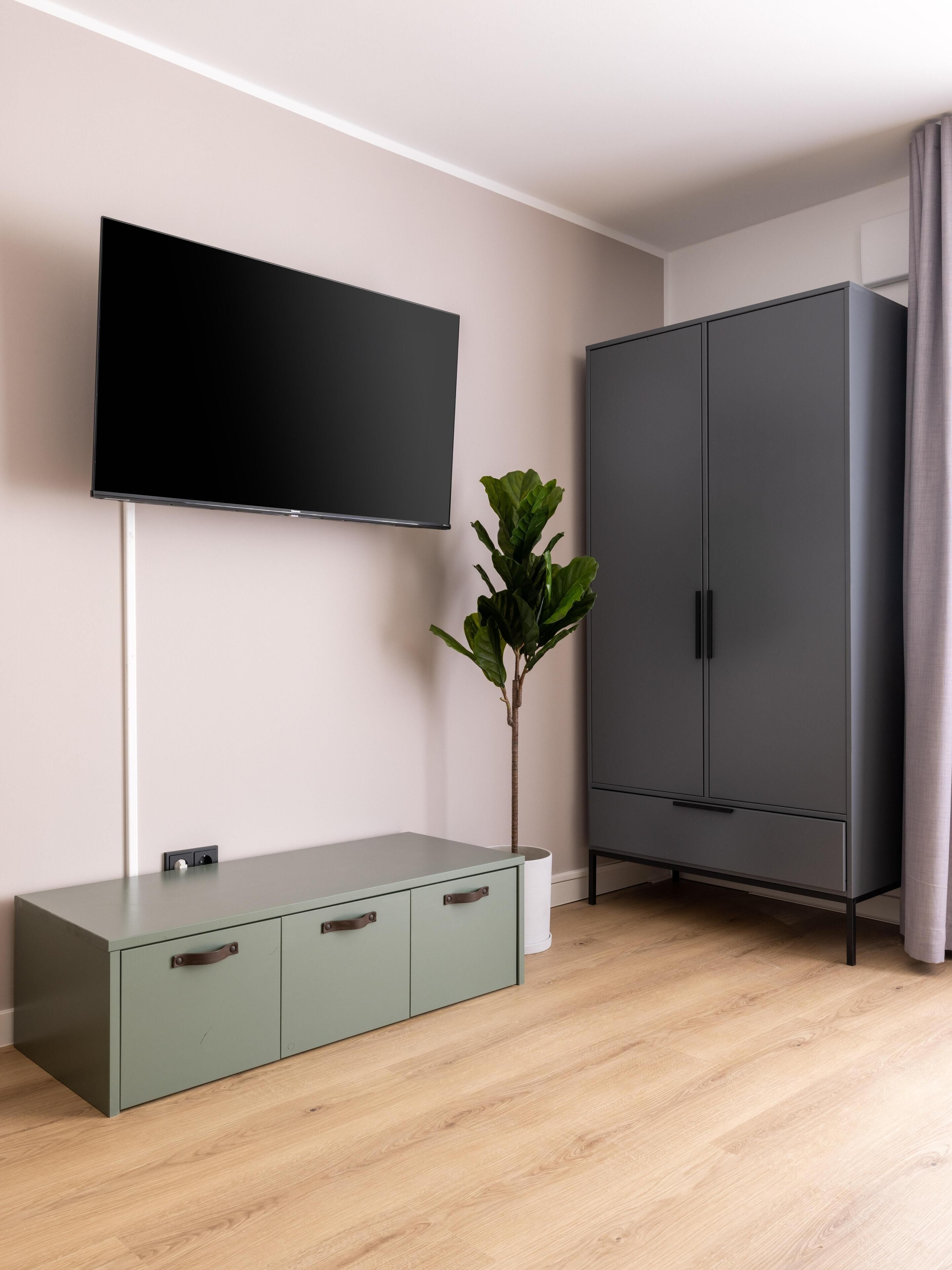 A modern room with a flat-screen TV mounted on the wall, a grey wardrobe, a green cabinet, and a potted plant.