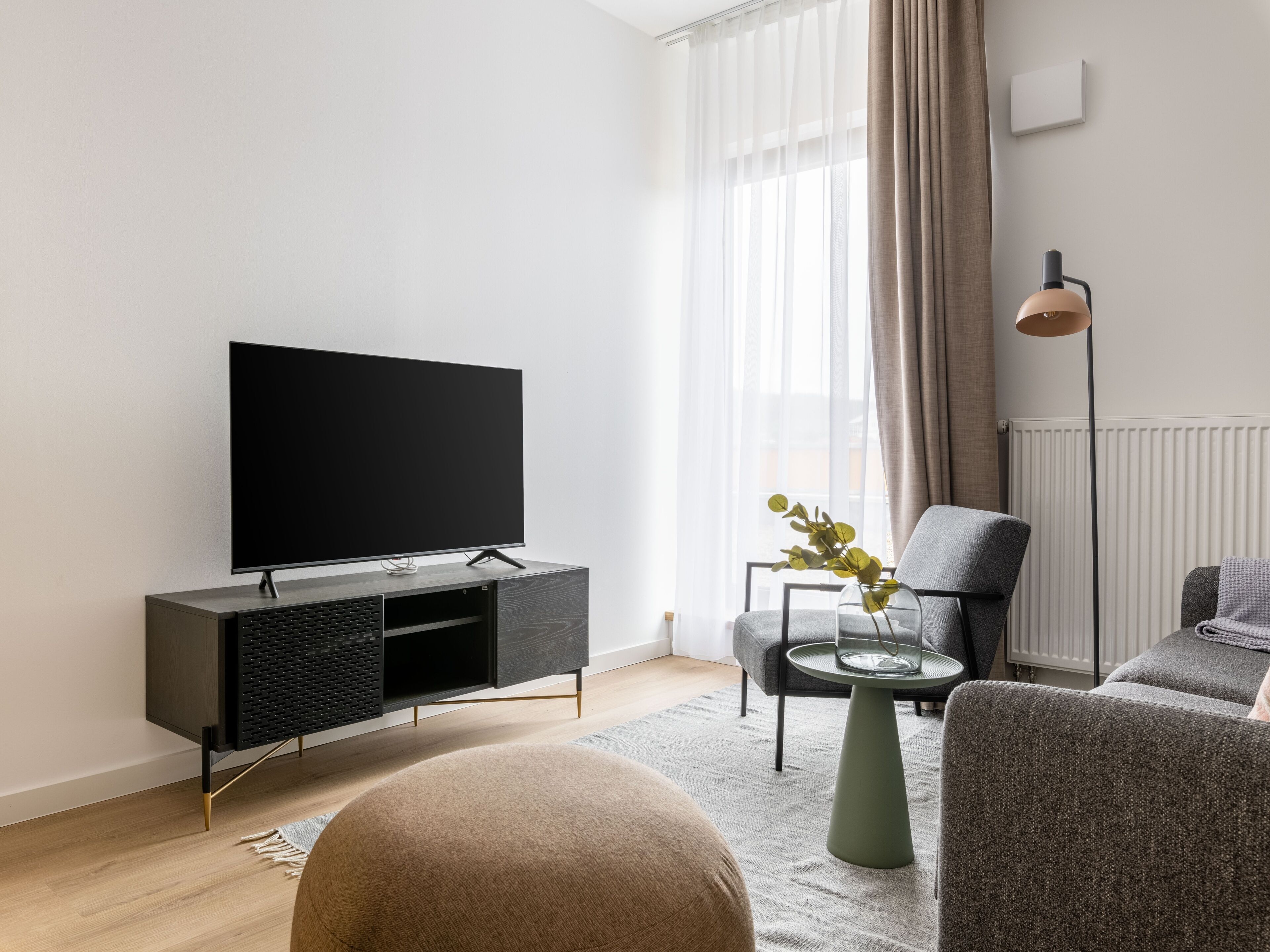 Un salon moderne avec un téléviseur à écran plat posé sur un meuble en bois foncé, un fauteuil gris, une table basse ronde ornée d’un vase de plantes vertes et un lampadaire.