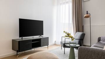 Un salon moderne avec un téléviseur à écran plat posé sur un meuble en bois foncé, un fauteuil gris, une table basse ronde ornée d’un vase de plantes vertes et un lampadaire.