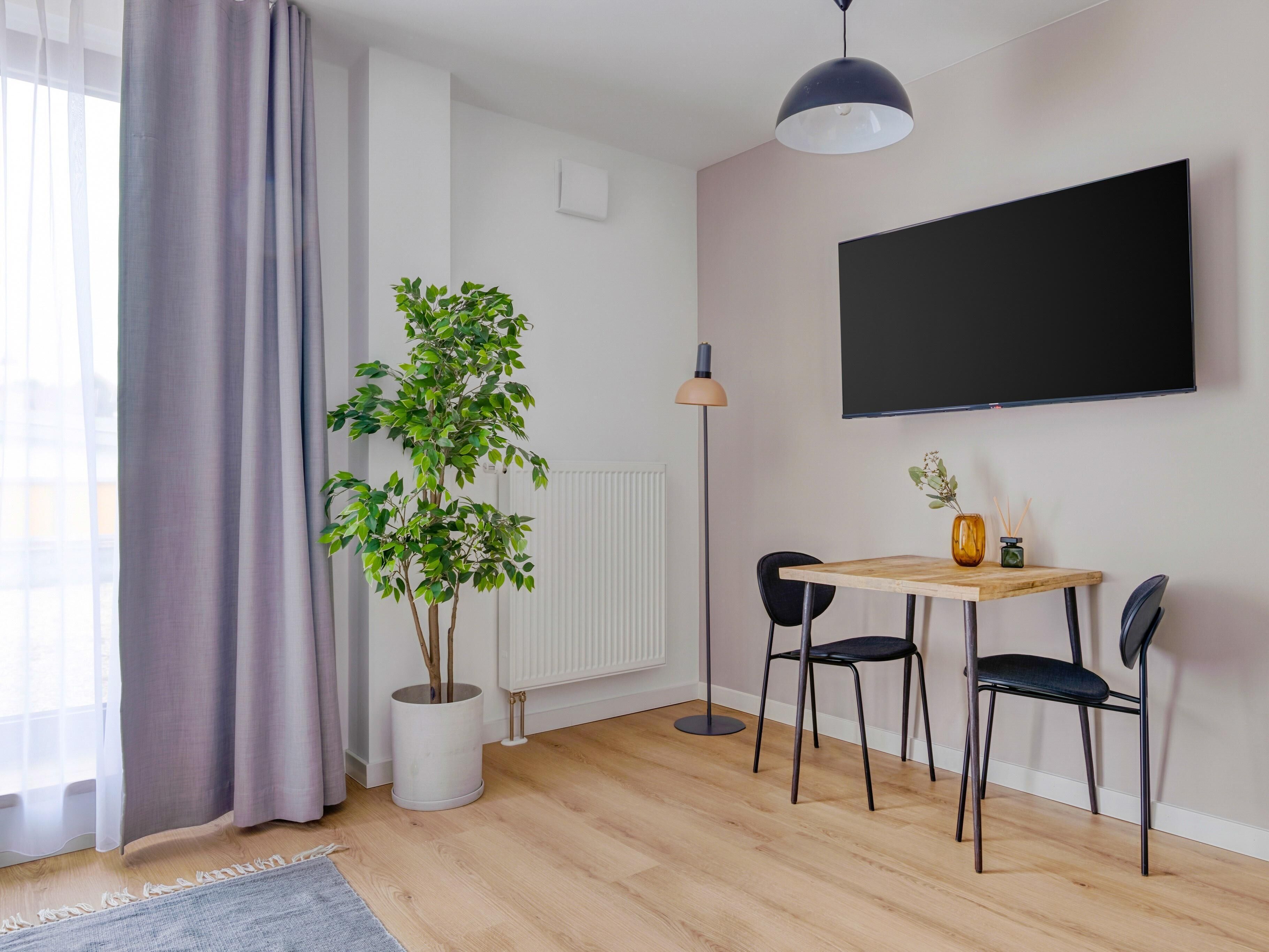 Una sala de estar moderna con suelo de madera, una mesa redonda, dos sillas negras, una planta en maceta, una lámpara de pie y un televisor grande montado en la pared.