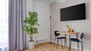 Una sala de estar moderna con suelo de madera, una mesa redonda, dos sillas negras, una planta en maceta, una lámpara de pie y un televisor grande montado en la pared.