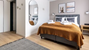 A modern hotel room with a large bed, a round mirror, and a view of the adjacent room.