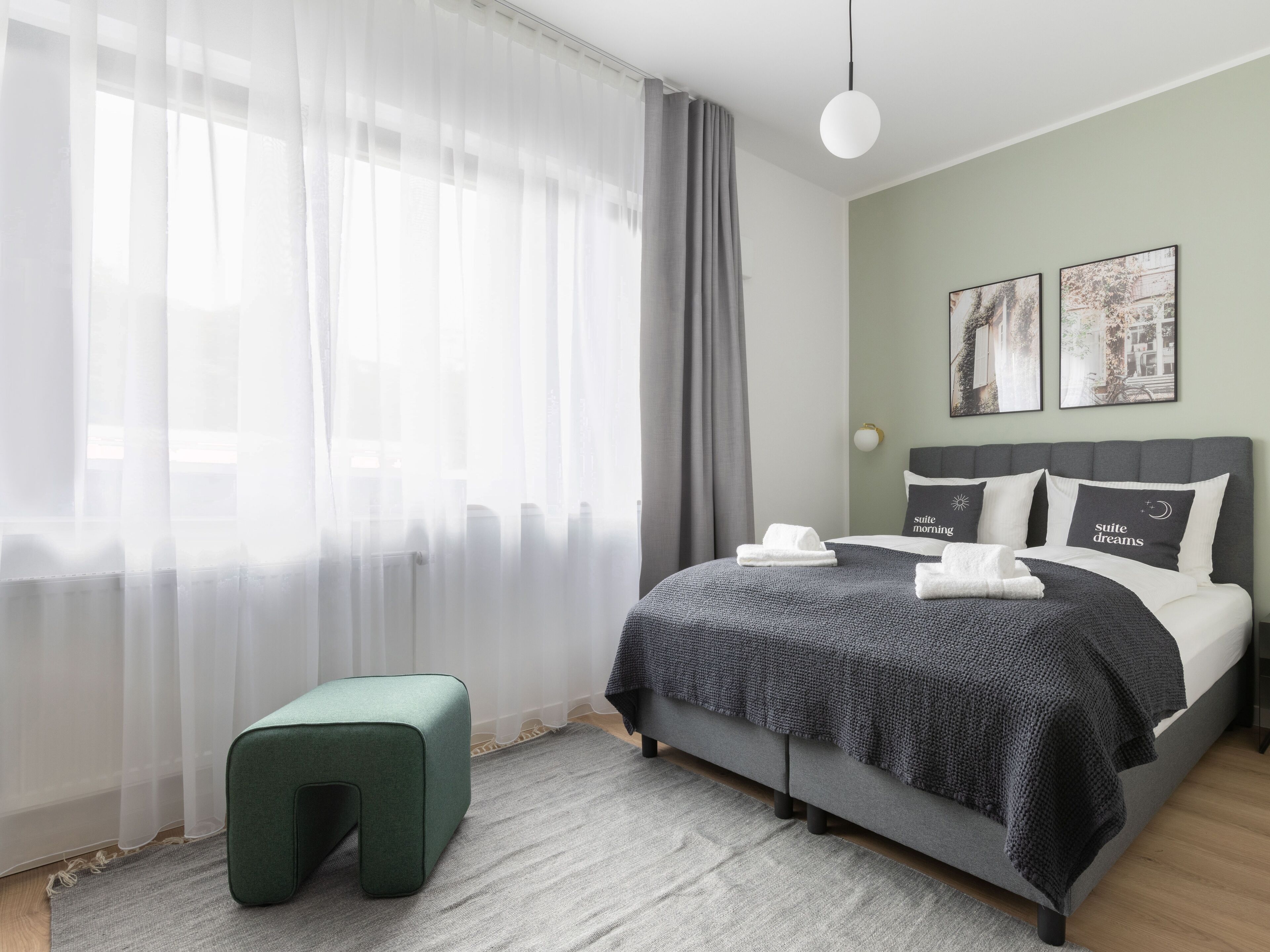 Une chambre moderne avec un lit, un pouf vert et une grande fenêtre ornée de rideaux légers.